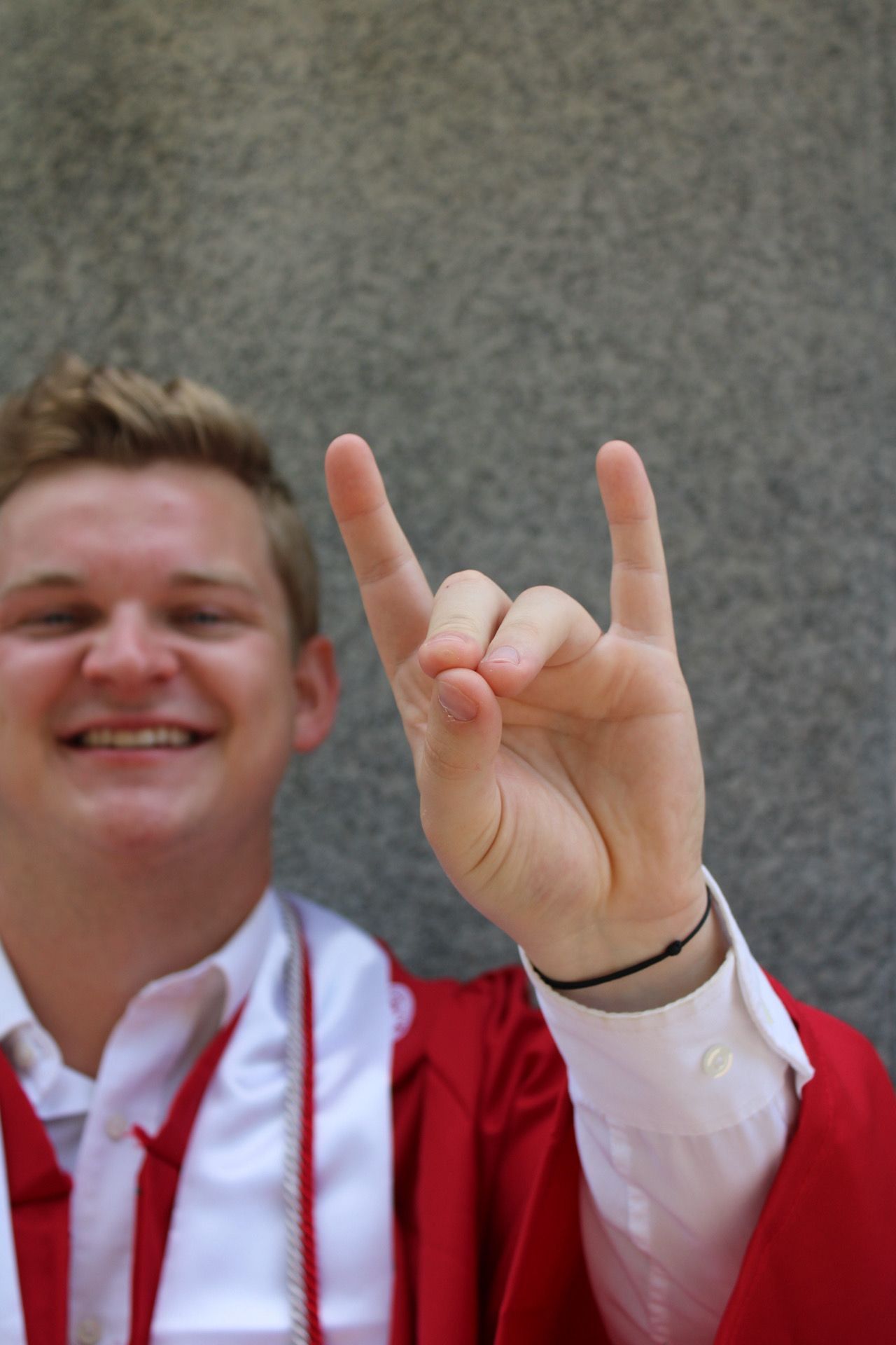 Someone in a red jacket makes a peace sign gesture against a grey wall.