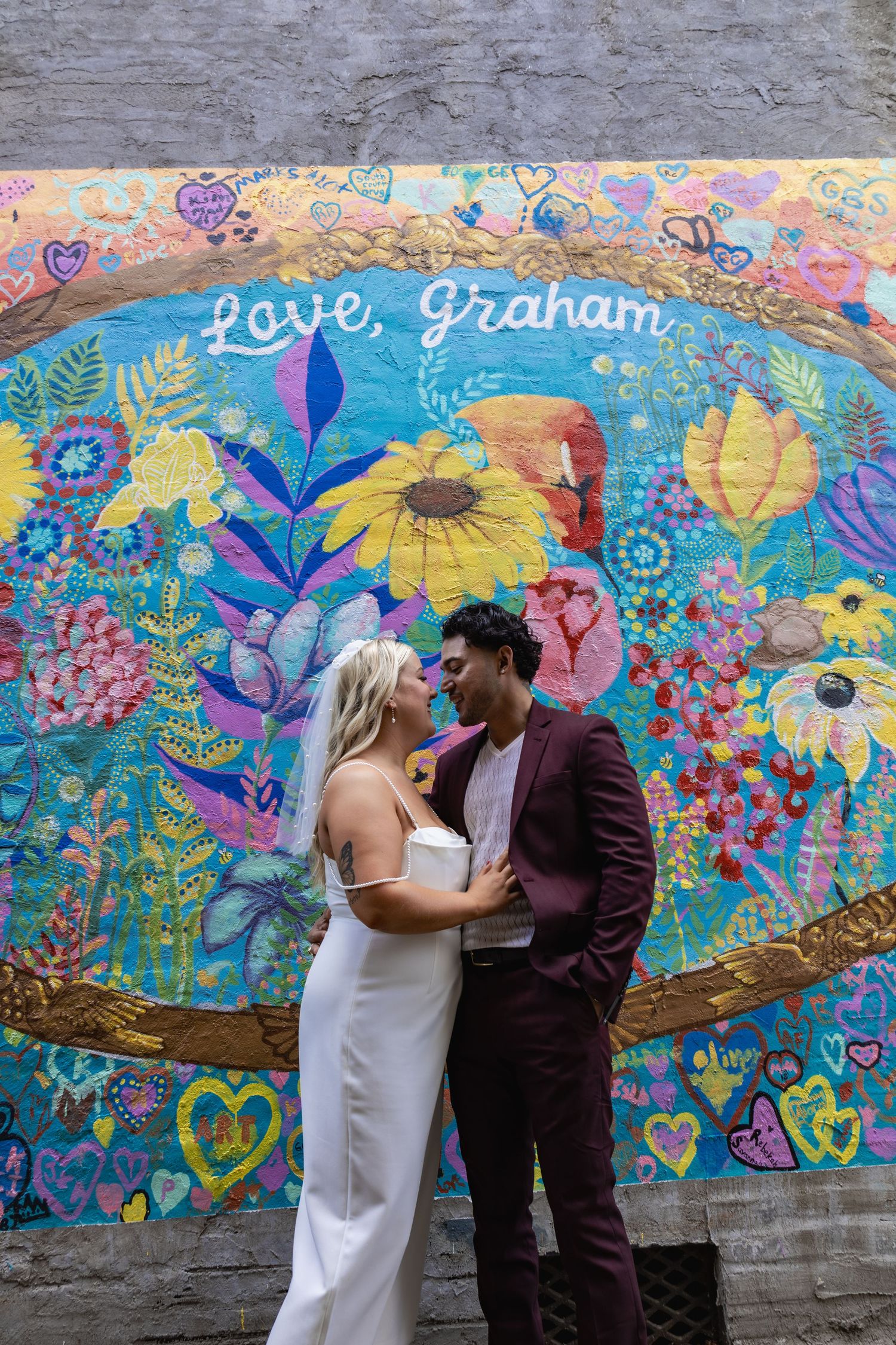 Couple embracing in front of vibrant floral street art mural with hearts and sunflowers.