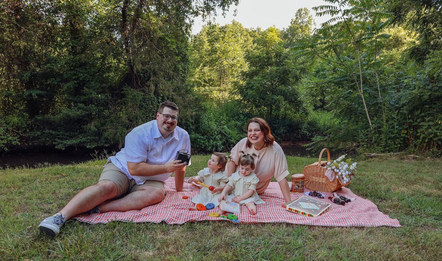 Picnickers relax on a red checkered blanket surrounded by trees in a park.