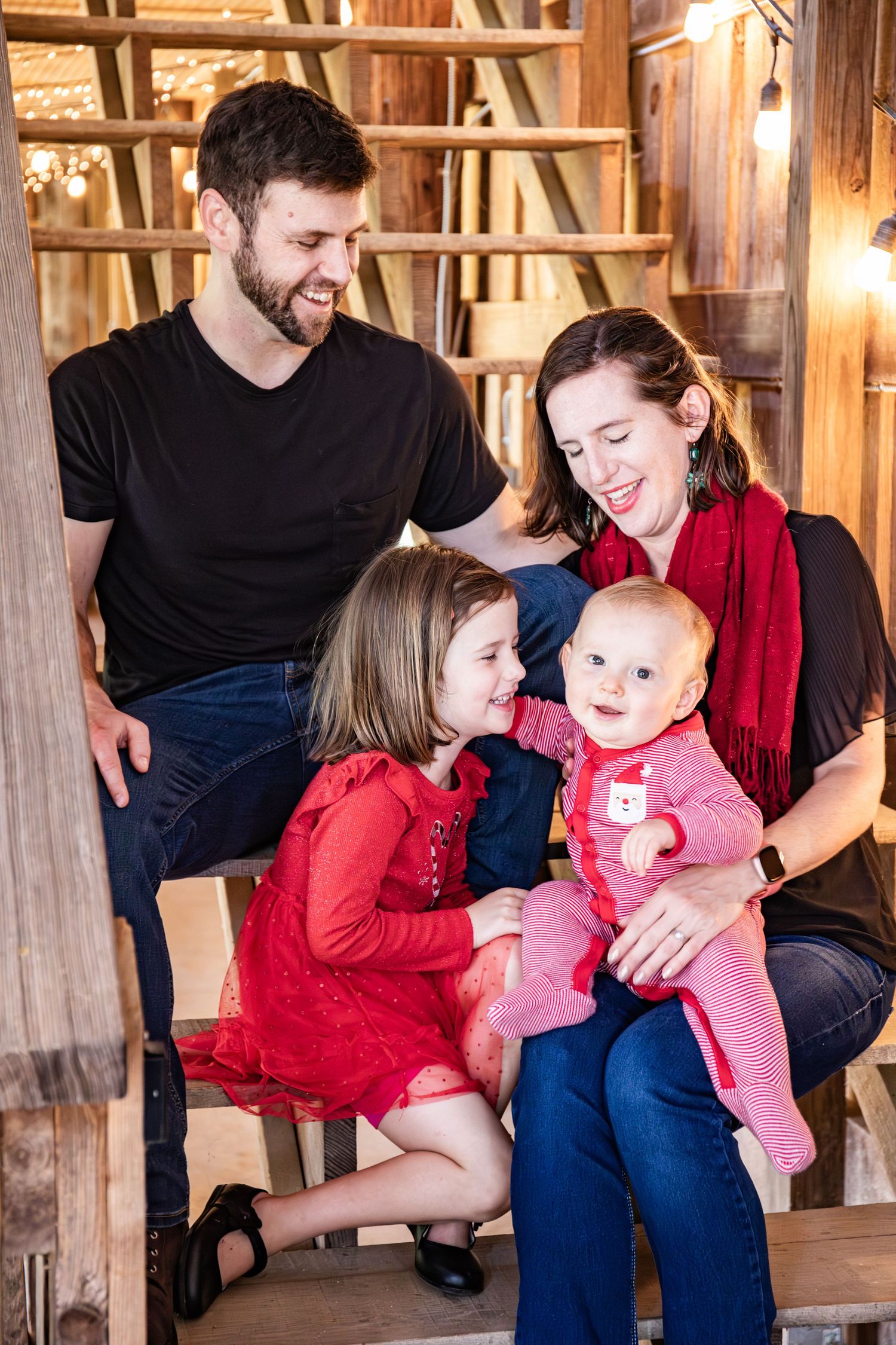 A family in red and black outfits shares tender moments together in a rustic wooden barn setting.