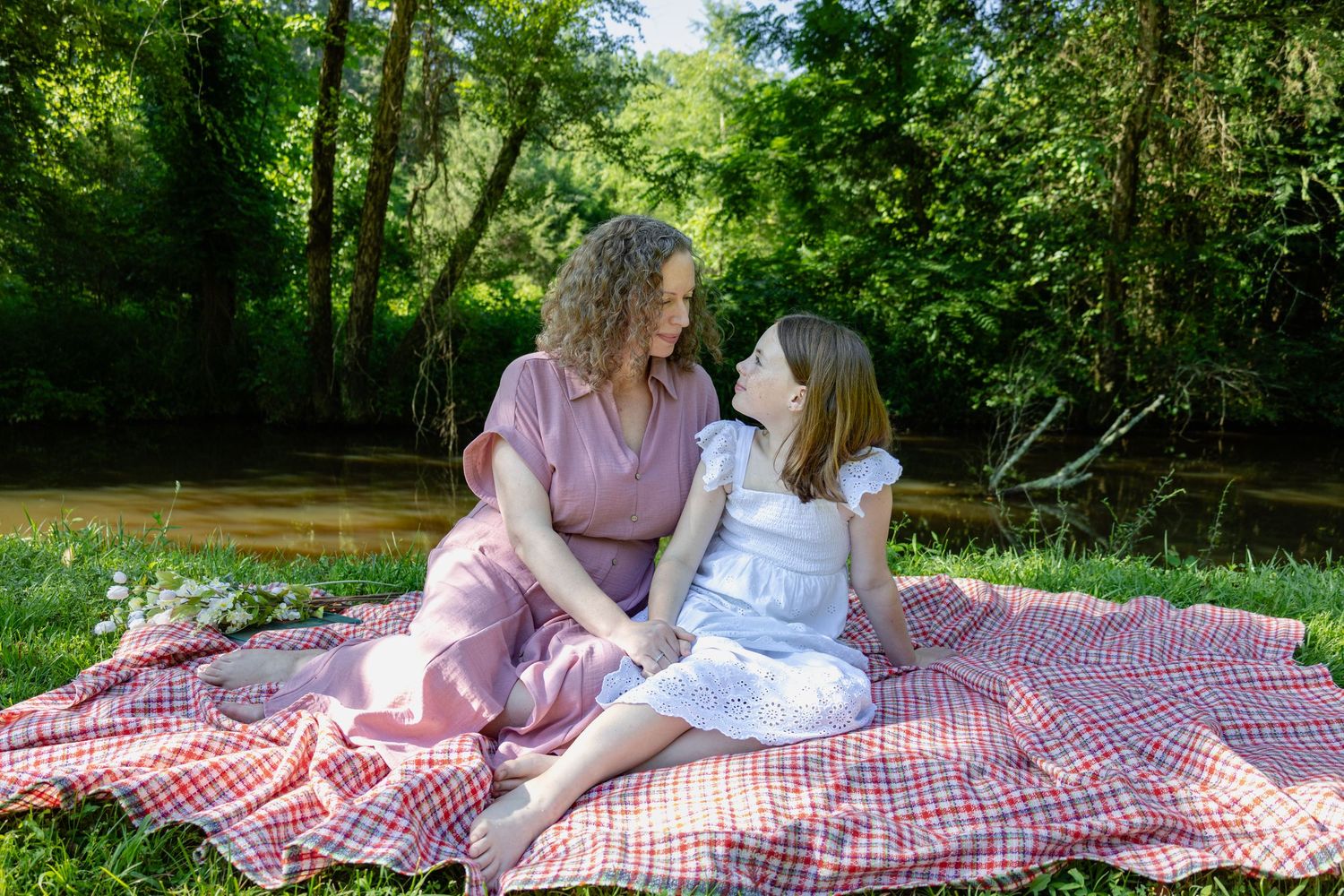 A group enjoys a relaxing summer picnic on a red and white checkered blanket in a lush green park setting.