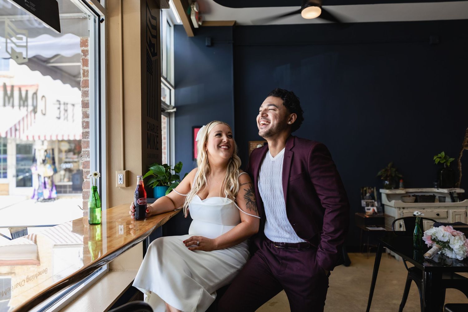 A couple laughs together while sitting by a cafe window in wedding attire.