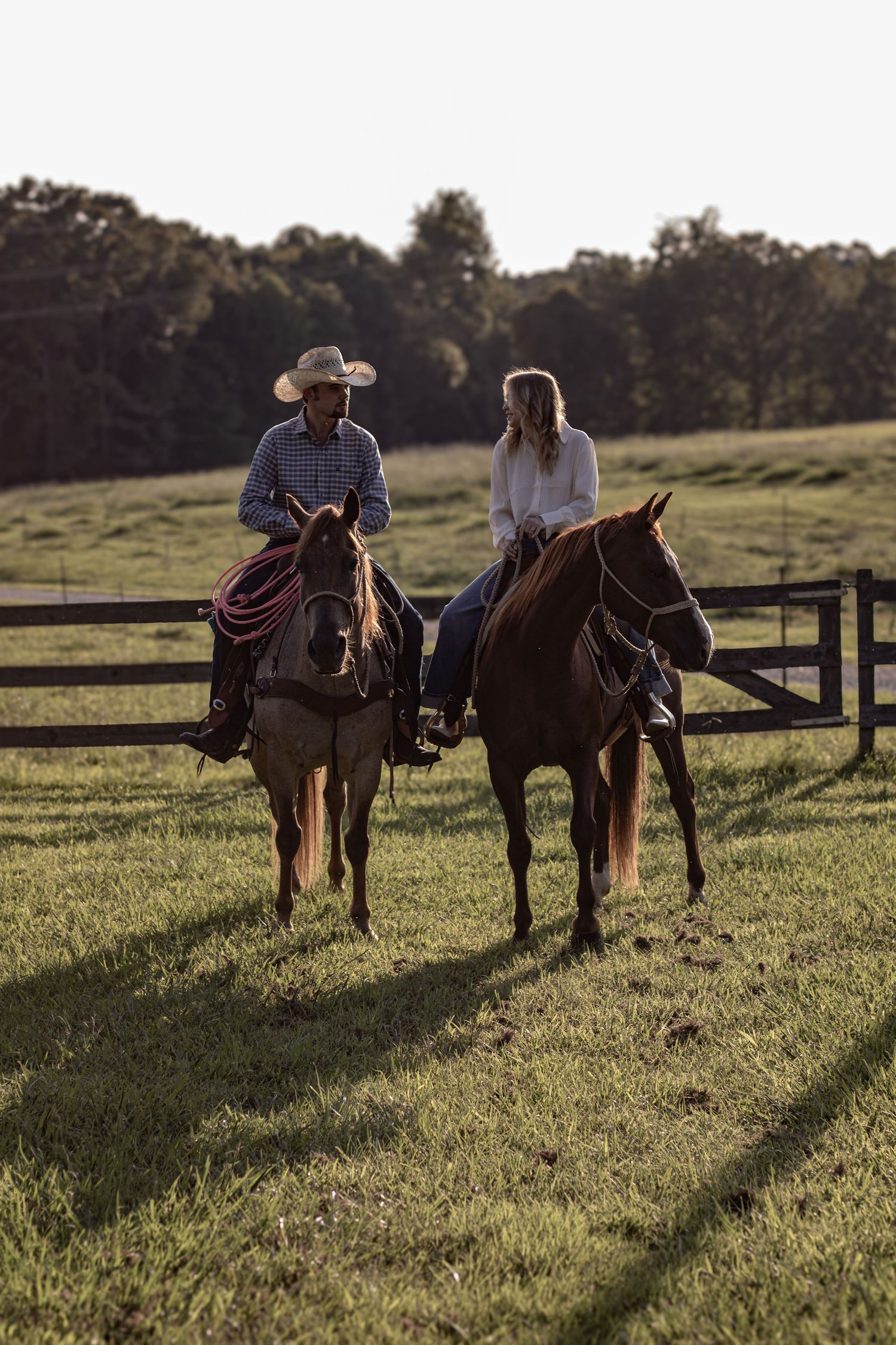 Two horses and riders resting along fence at sunset.