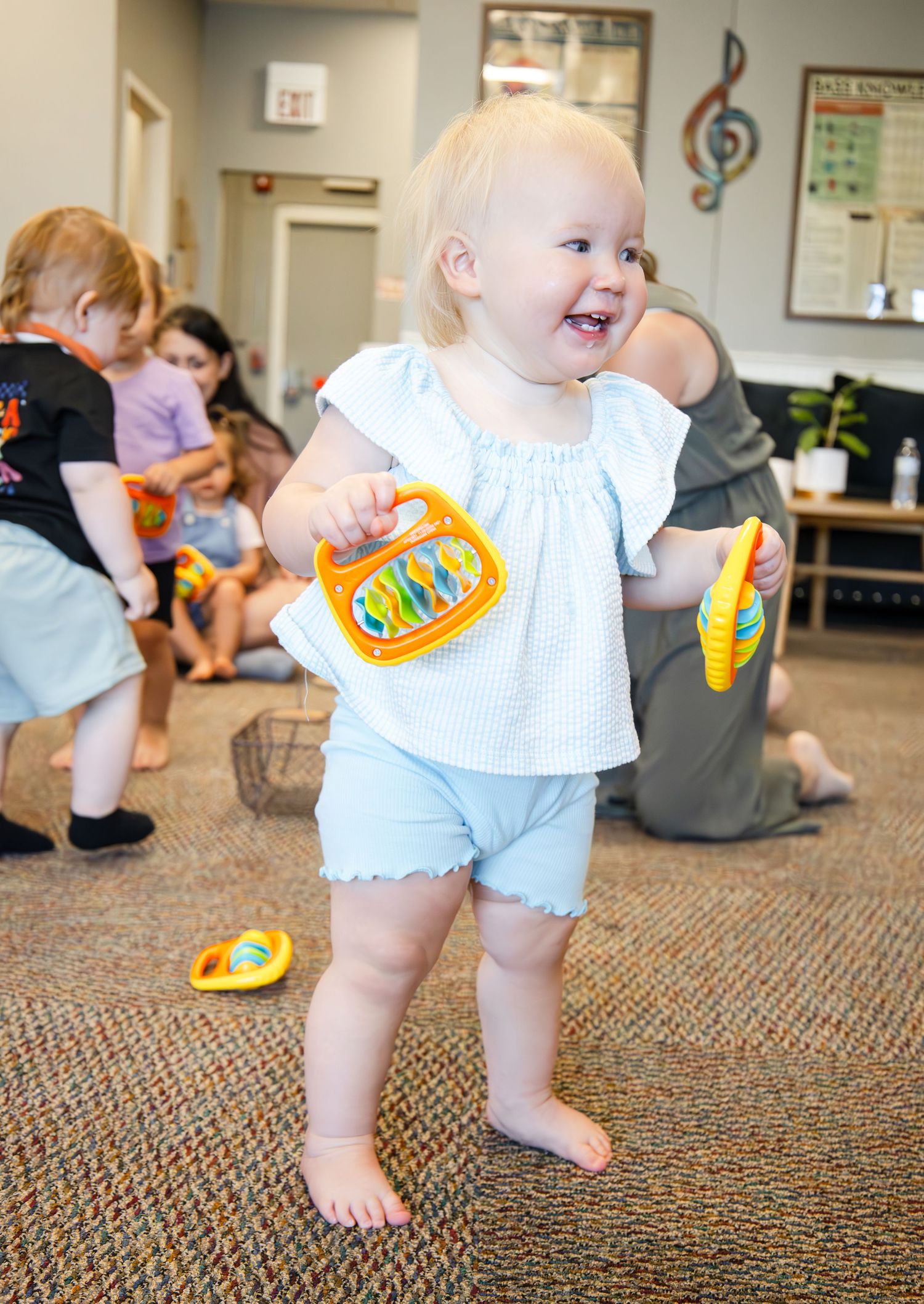 A toddler in light blue shorts and white top laughs while holding yellow toys during an indoor music class.