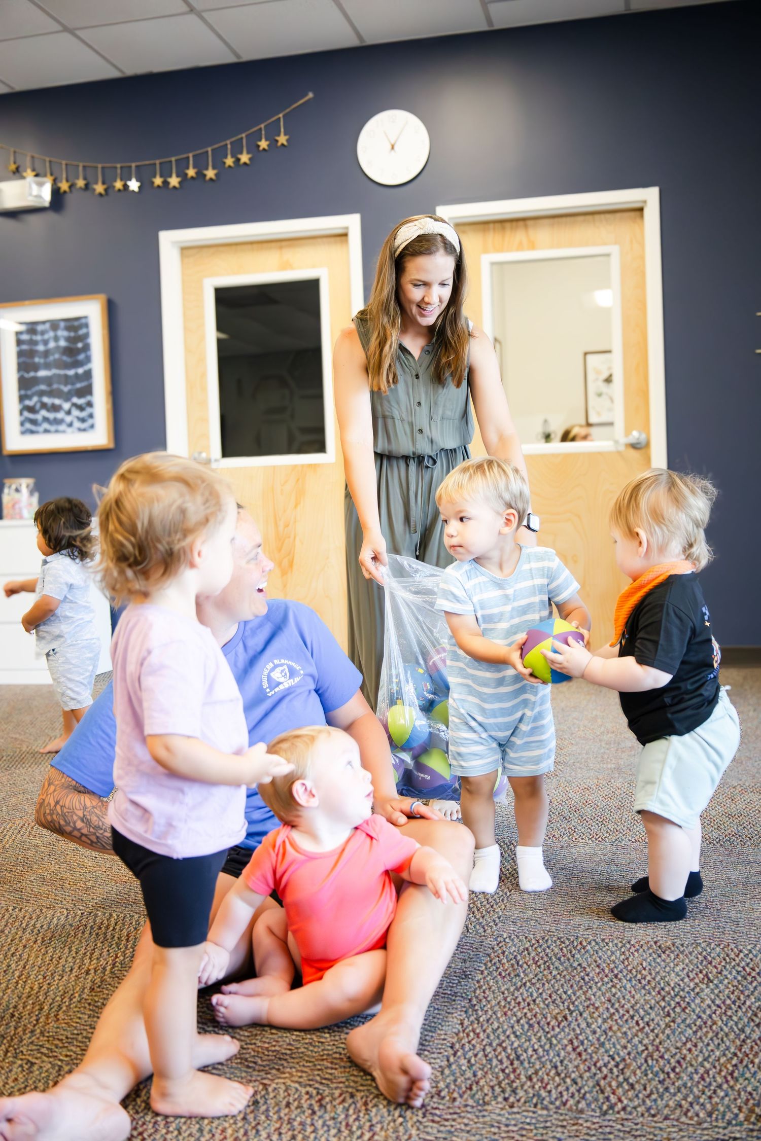A group of toddlers enjoy playtime with their caregiver on a carpeted floor with string lights and a clock on the wall.