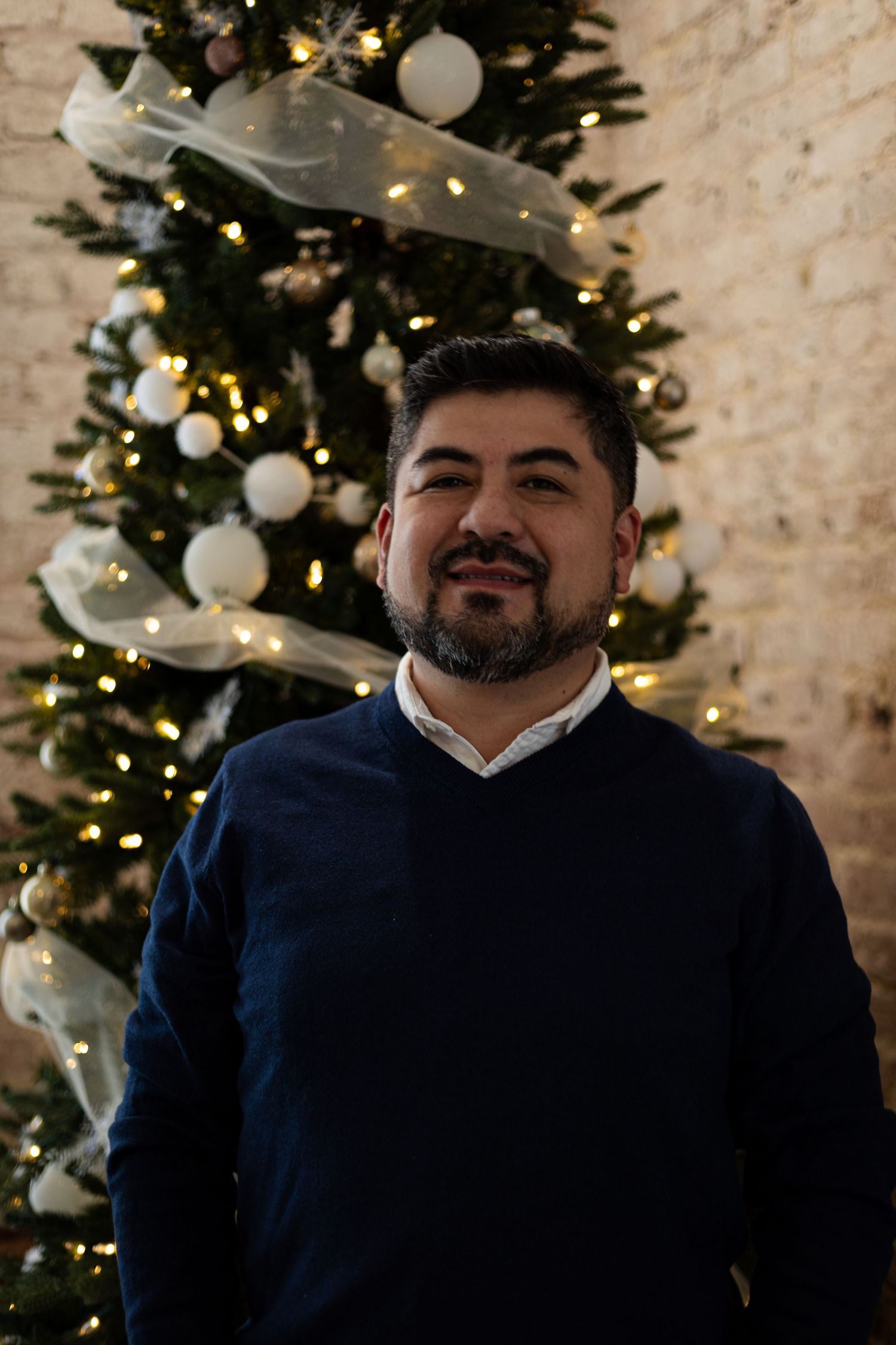 Man wearing navy suit jacket poses in front of decorated Christmas tree with twinkling lights and stone wall backdrop.