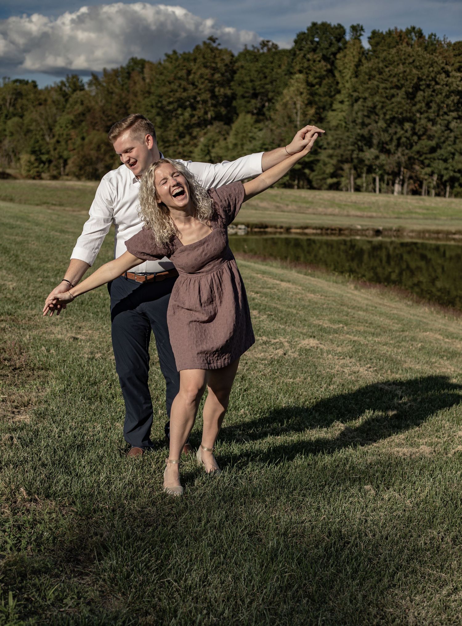 A couple playfully spins together on a grassy hillside near a lake on a cloudy autumn afternoon.