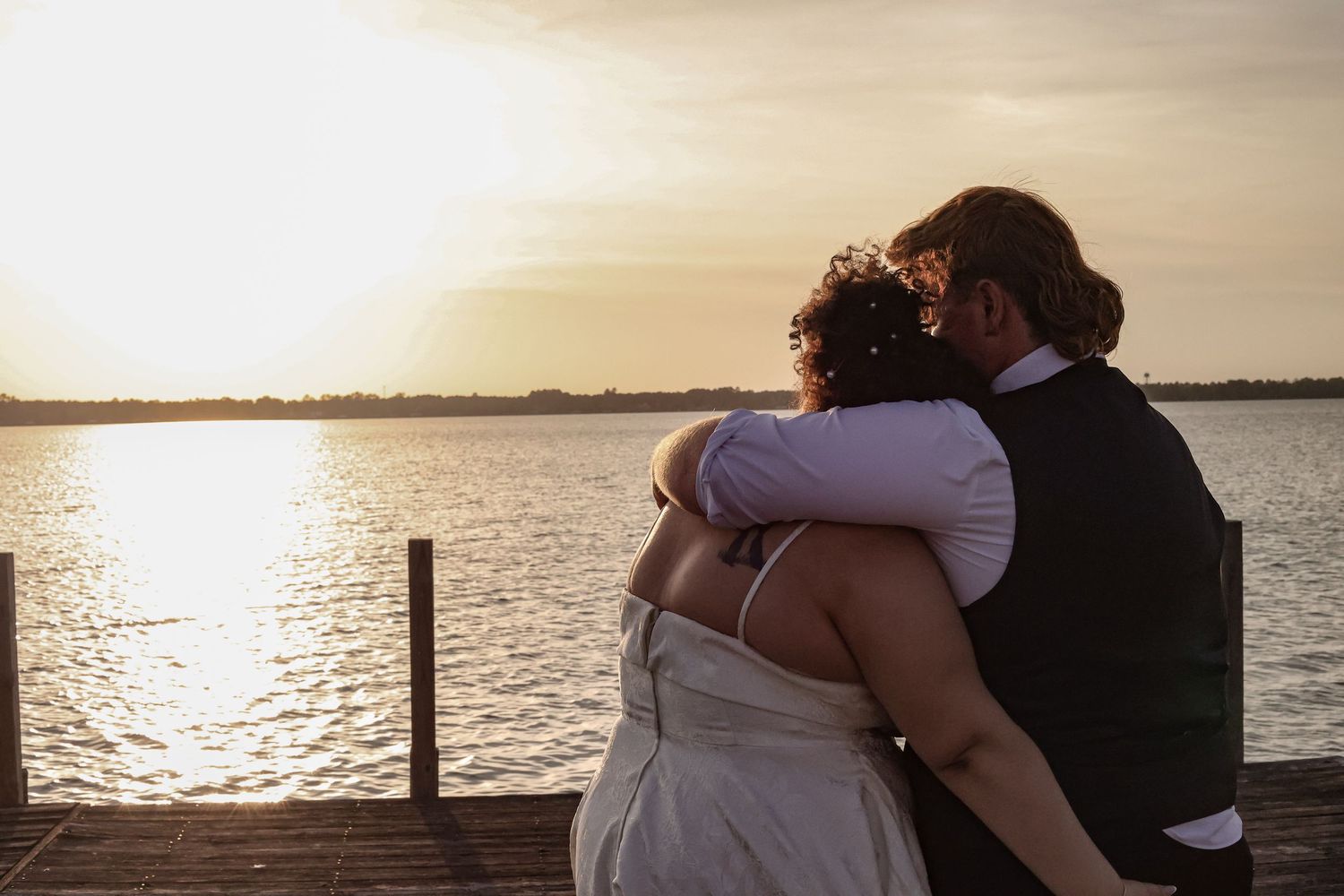 A couple embraces while watching the sunset over a lake from a wooden dock.
