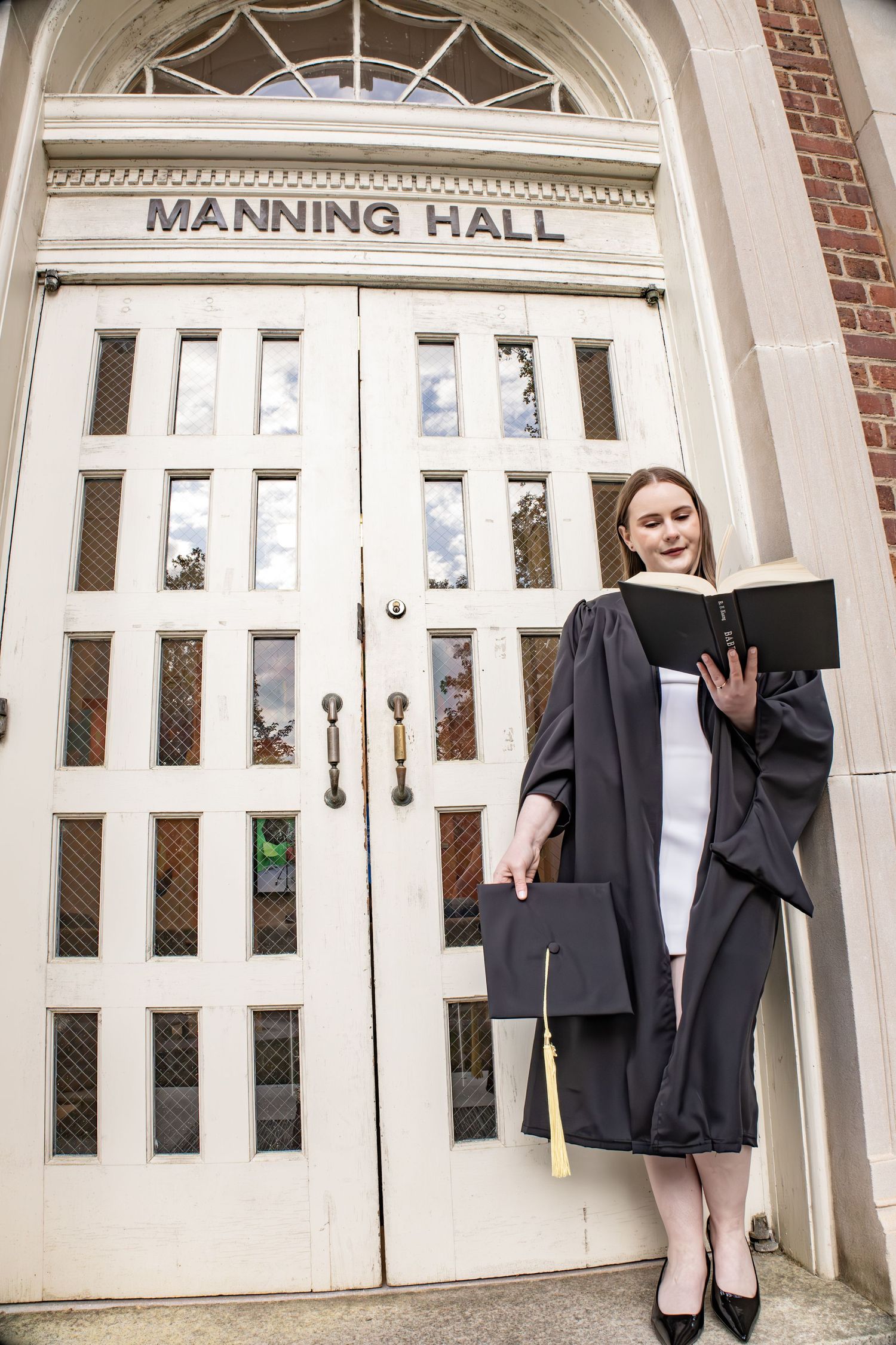 A graduate poses in academic regalia in front of Manning Hall's classic architectural entrance.