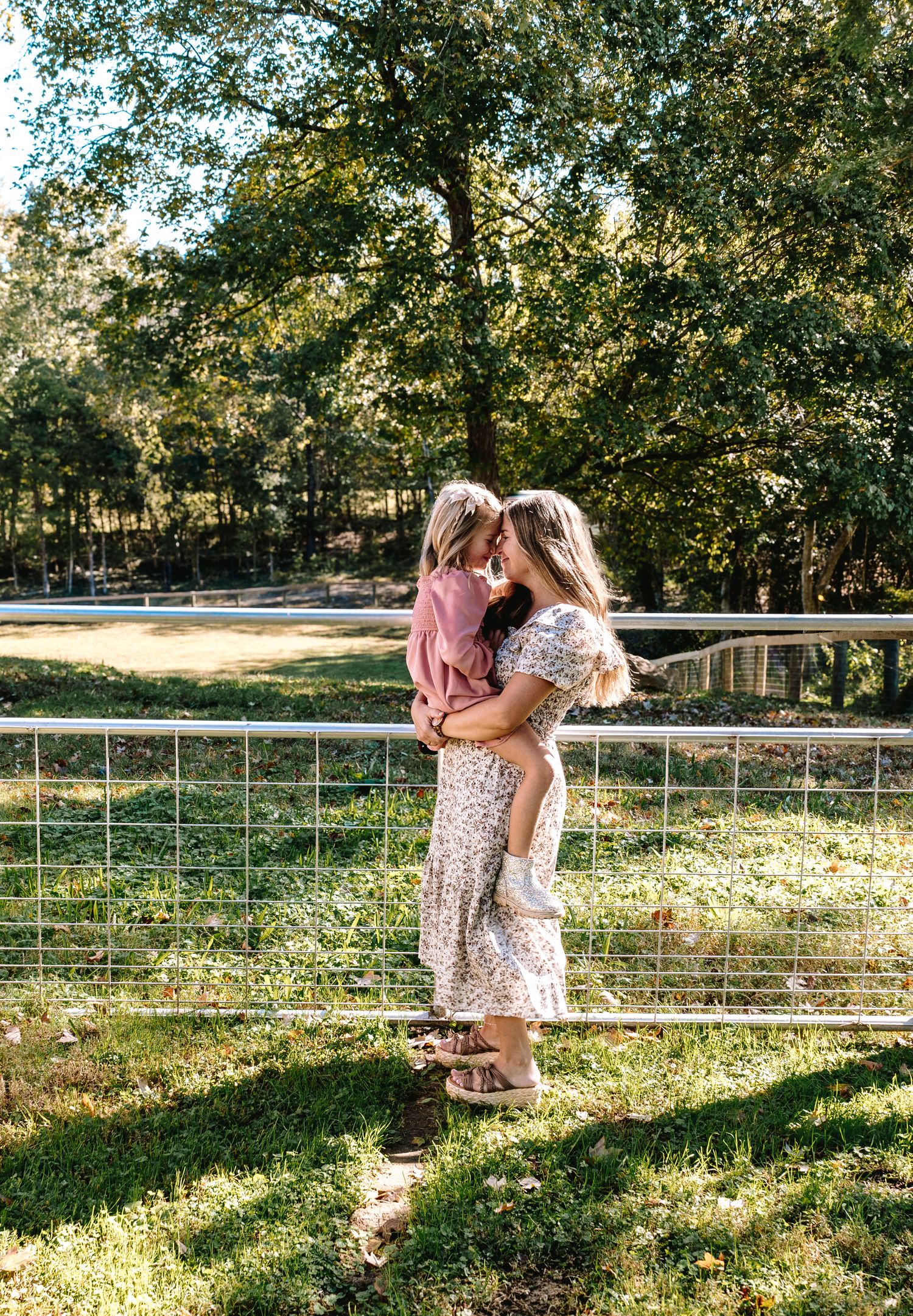 Mother and child embrace in sunlit outdoor setting with rustic fence and trees in background.