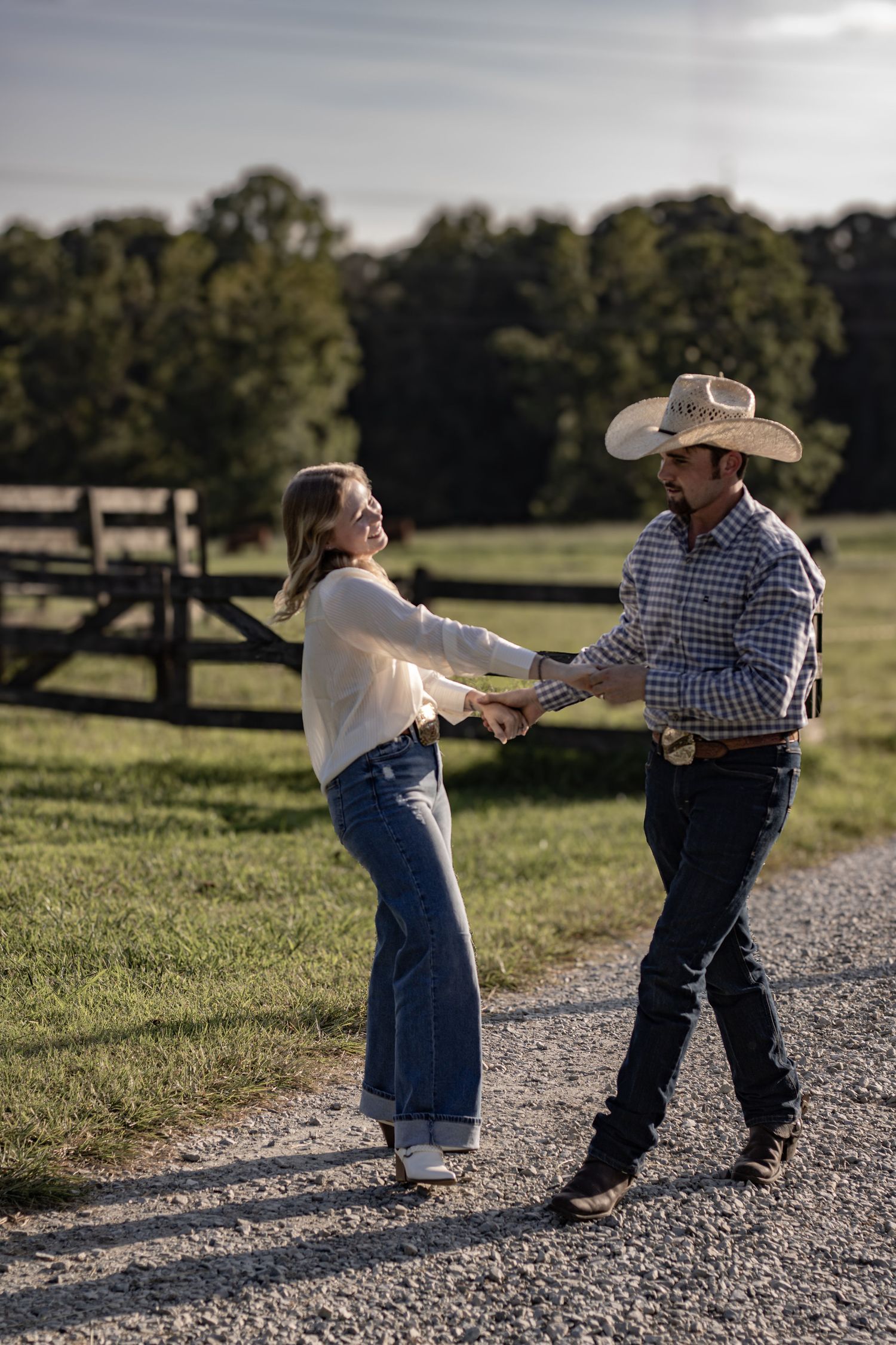 A playful couple dancing on a gravel path with rustic fencing in the background.