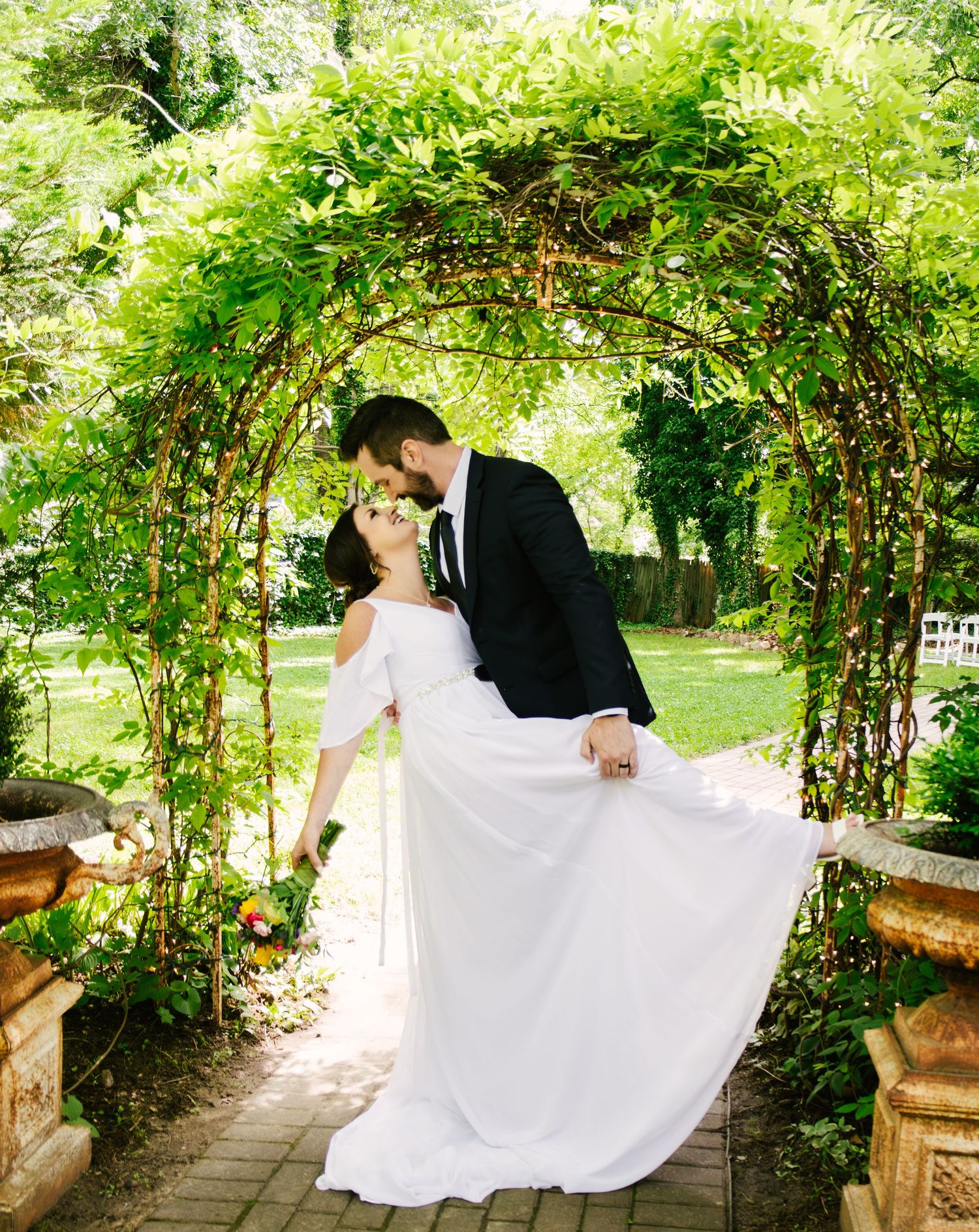 A newlywed couple shares a romantic moment under a lush green garden arch on a brick pathway in a garden setting.