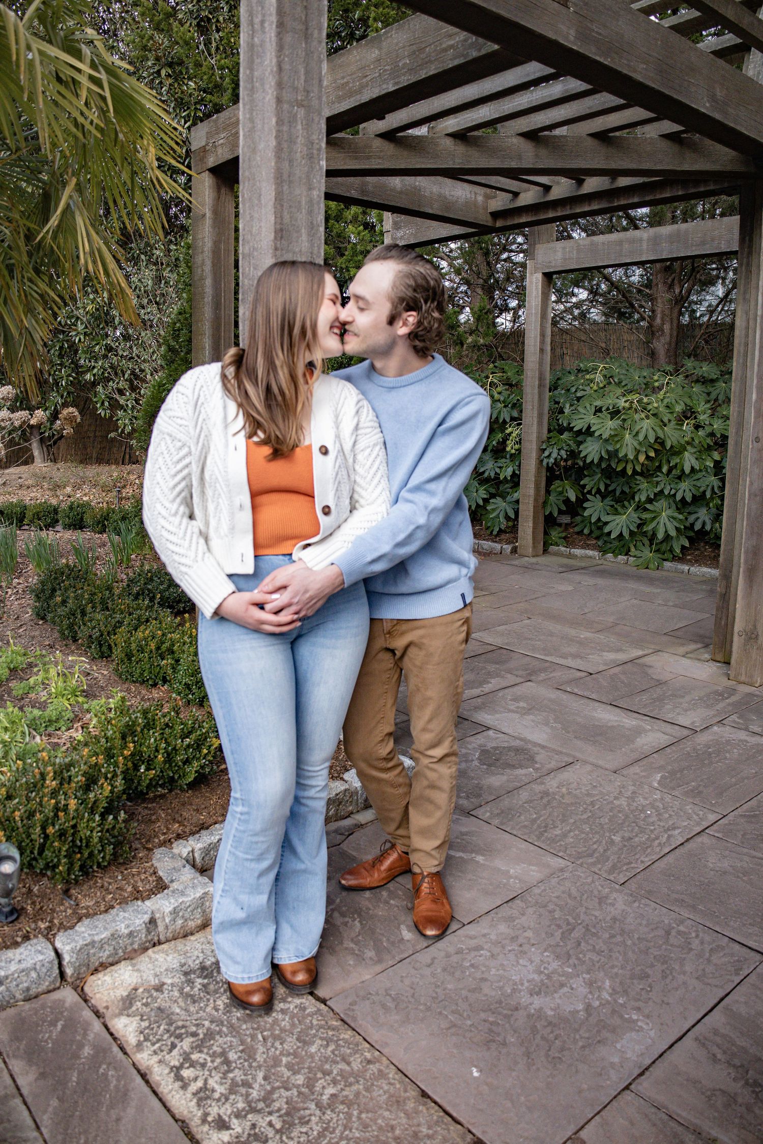 Couple embraces under wooden pergola in garden setting.