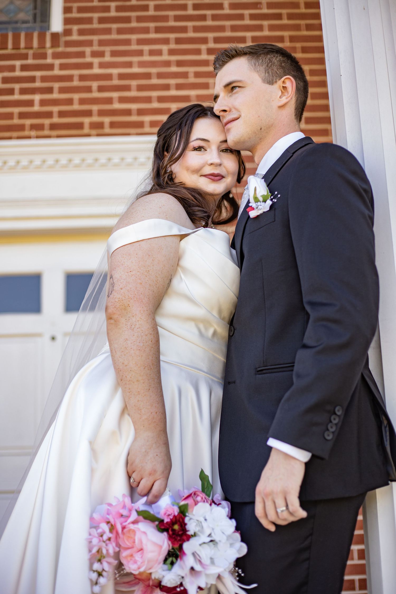 Bride in white off-shoulder gown holding pink bouquet leans on groom in navy suit near brick building.