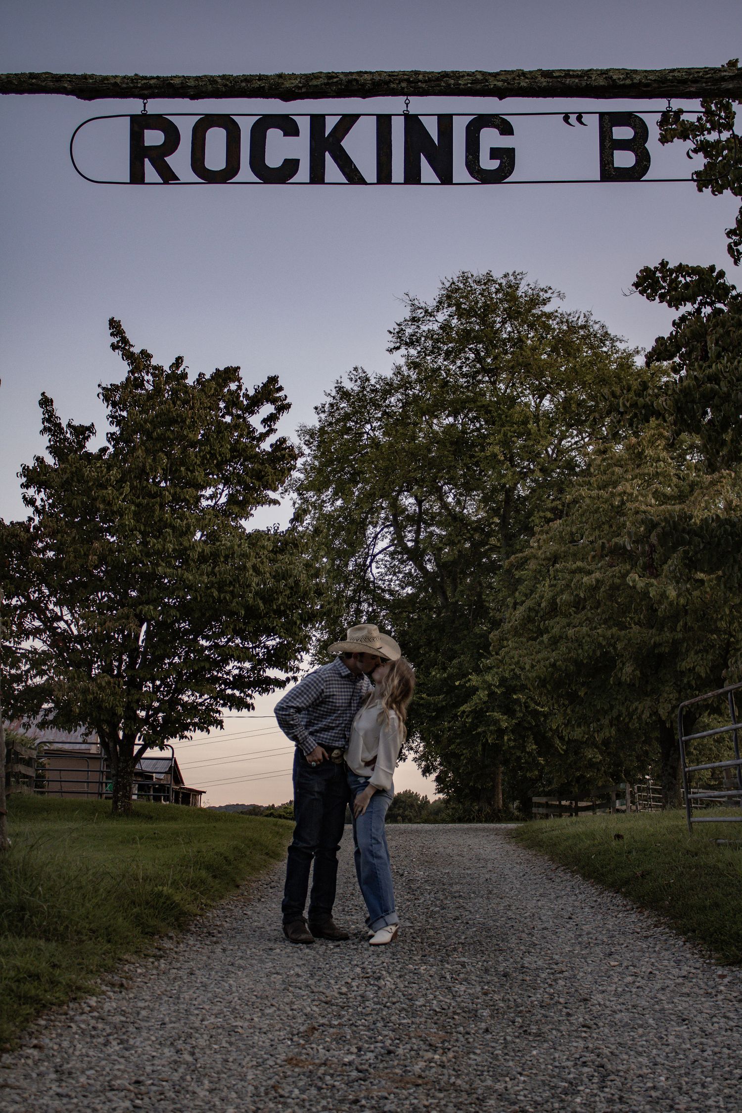 A couple embraces under a 'Rocking B' ranch sign on a gravel path at dusk.