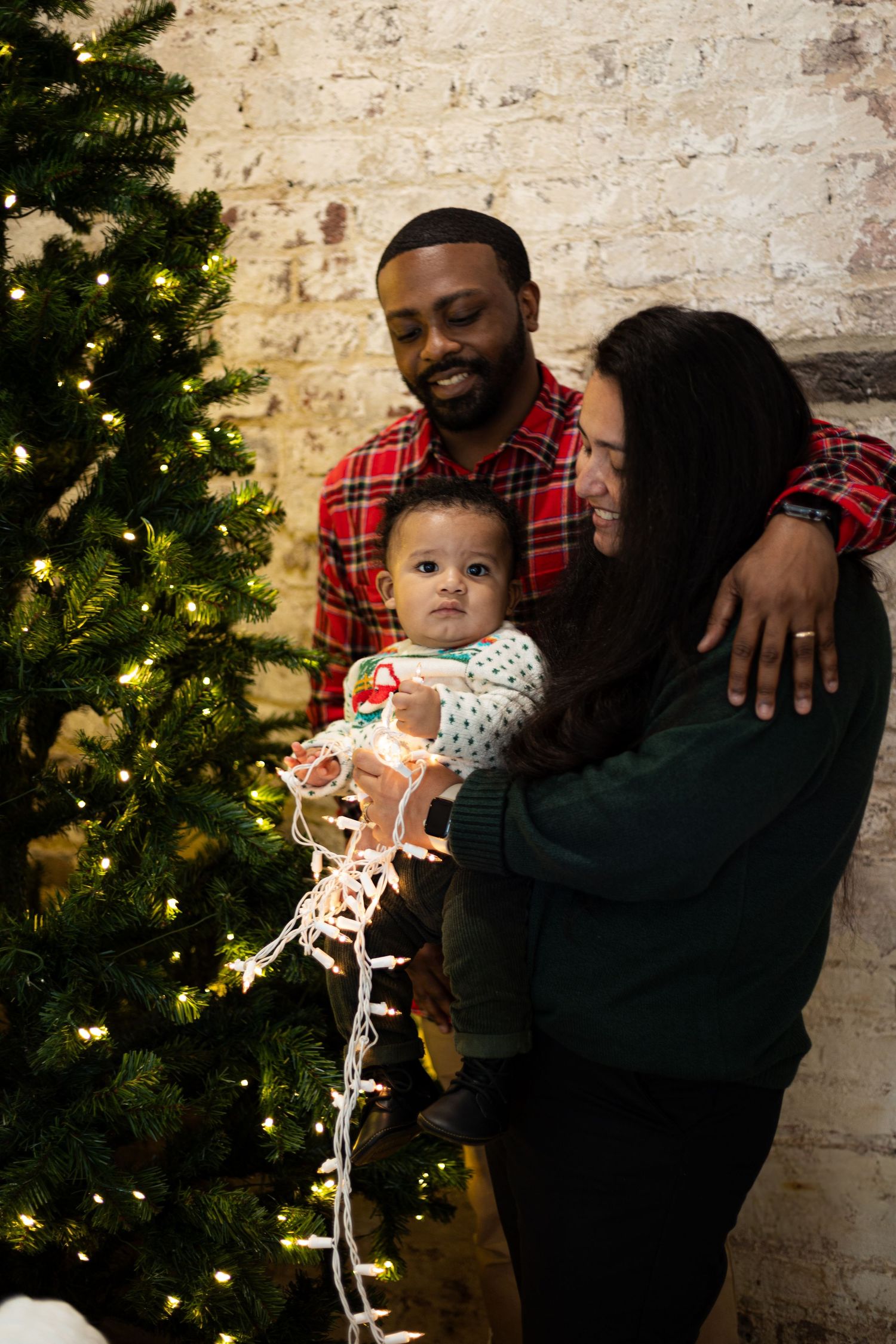 Playful family moment captured by Christmas tree with string lights and exposed brick wall.