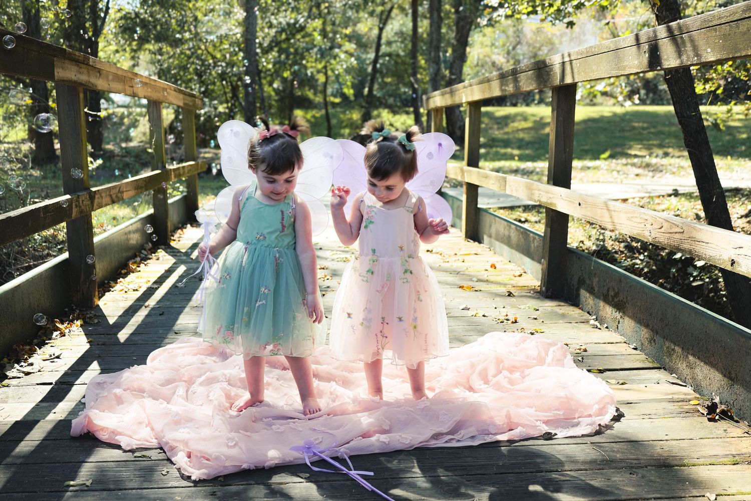 Two small children in pastel dresses stand together on a wooden bridge with a flowing pink dress train behind them.