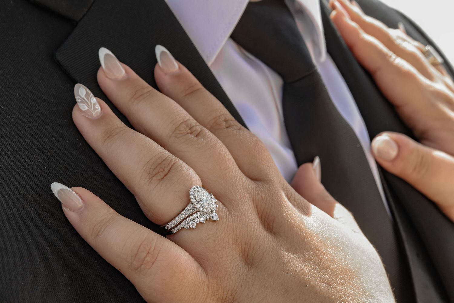 Close-up of bridal hand wearing silver engagement and wedding rings with sparkly manicured nails against suit.