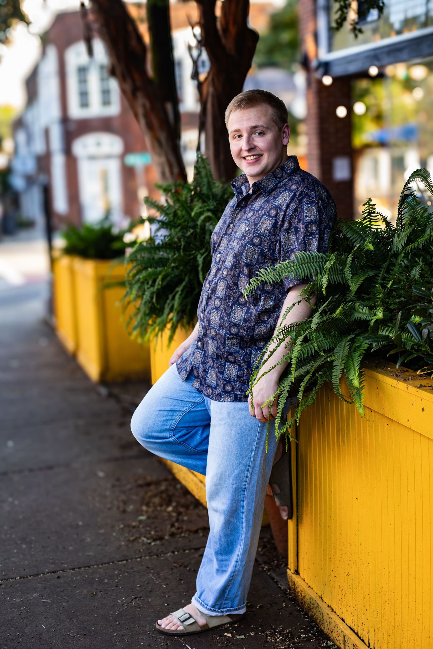 Person wearing navy patterned shirt and jeans sits casually on bright yellow planter box along sidewalk.