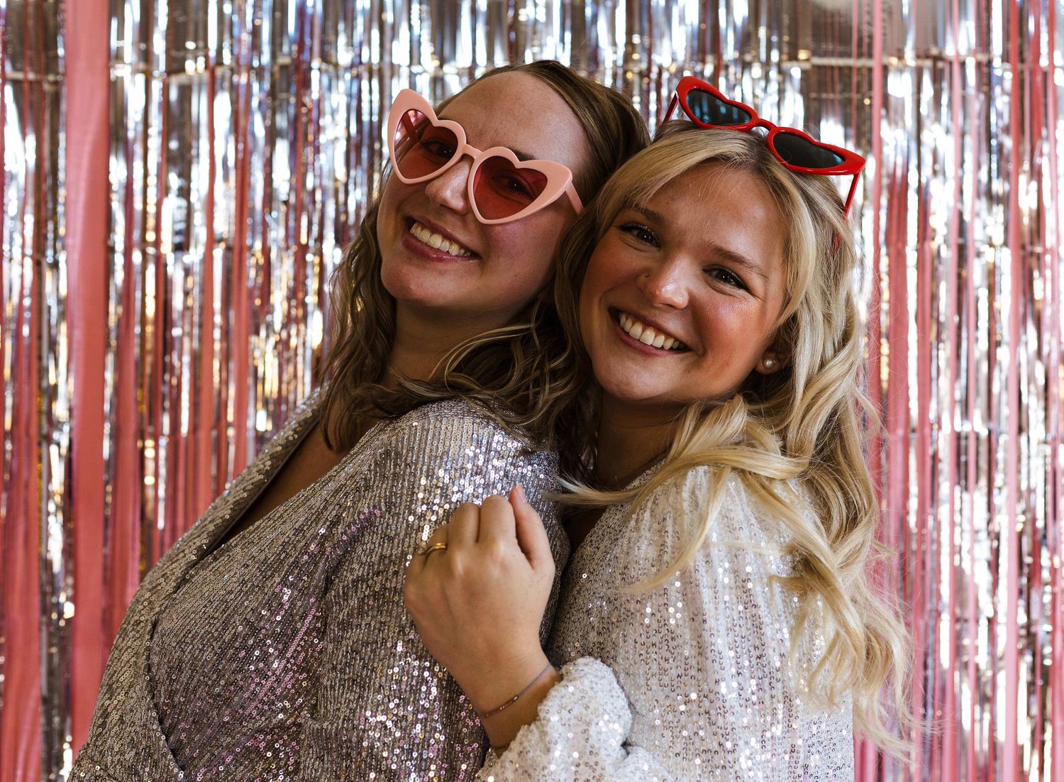 Two friends in sparkly outfits laughing together against a pink metallic fringe backdrop.