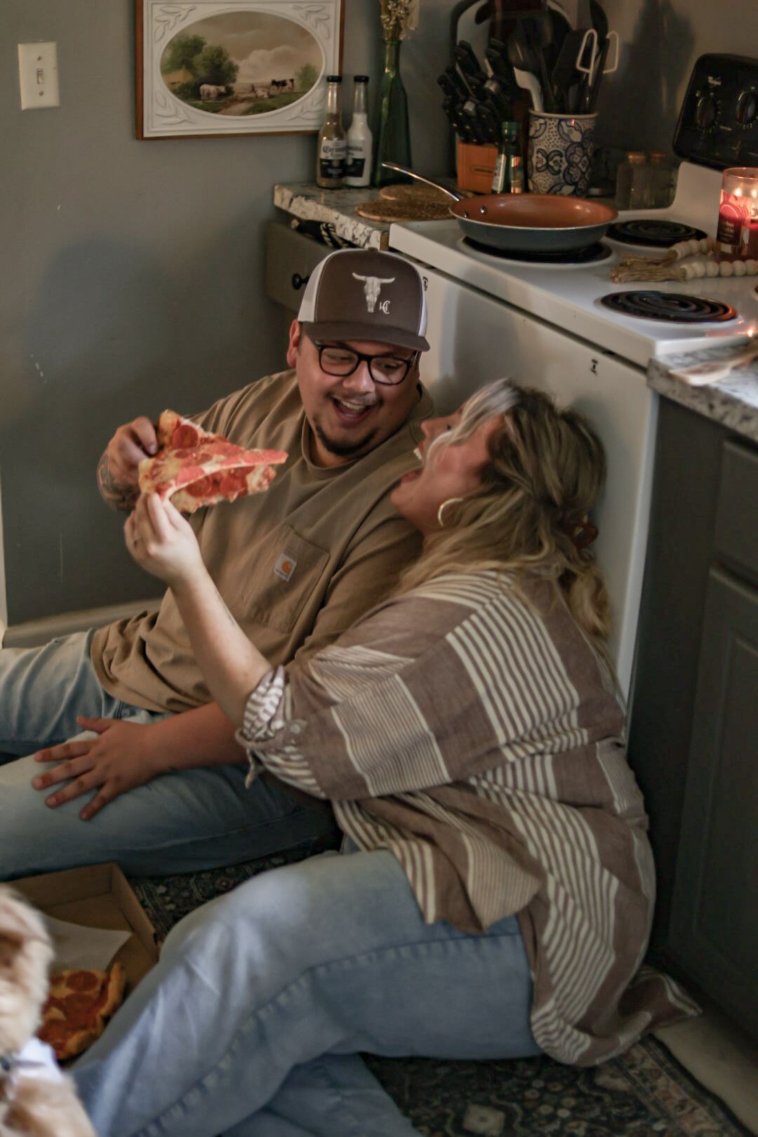 Two people laughing while sharing pizza on the kitchen floor.