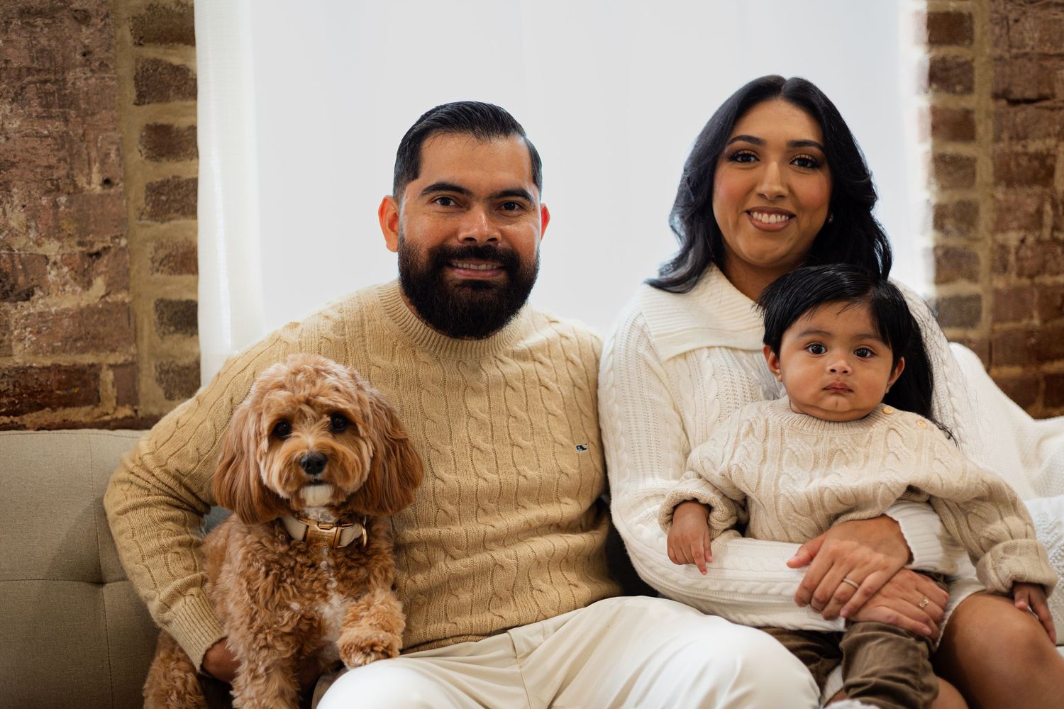 Cozy family portrait with small dog on brick background wearing matching cream sweaters.