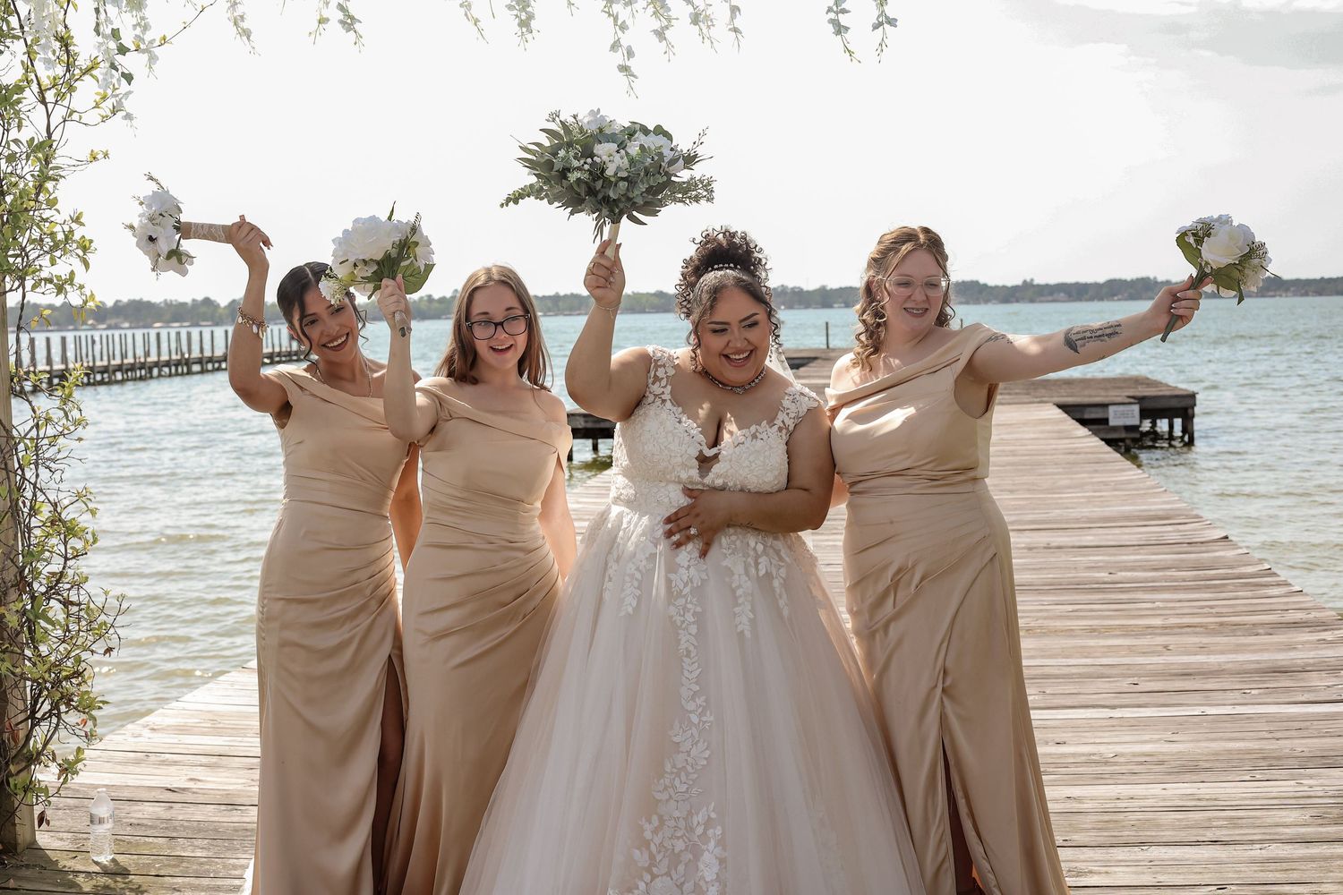 Bridesmaids in beige dresses celebrate joyfully on lakeside dock.