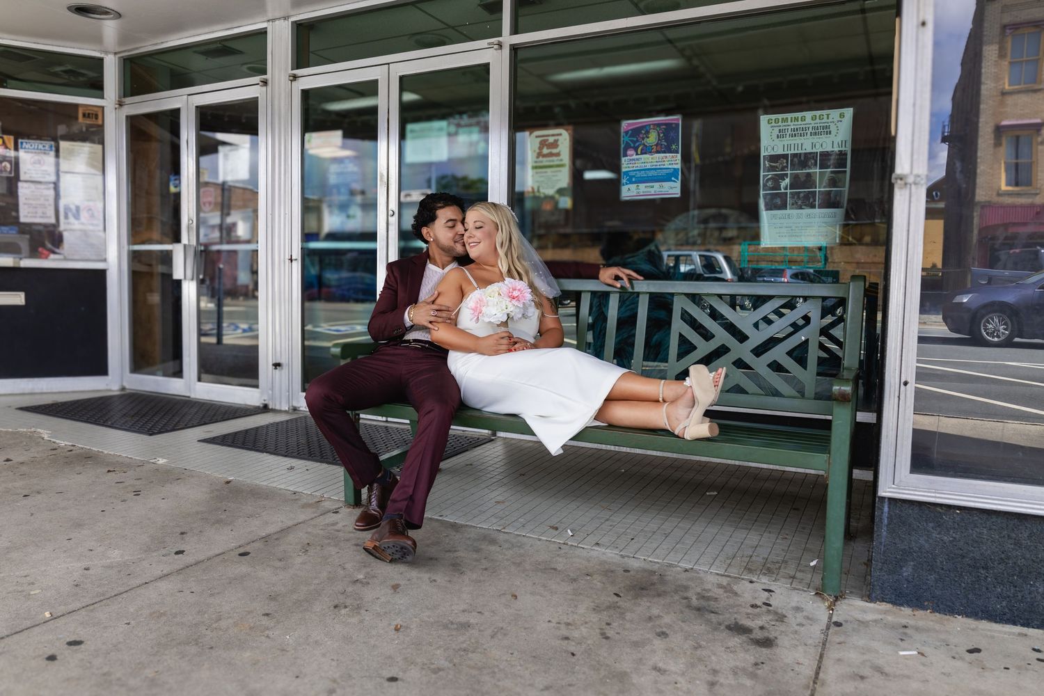 A couple in wedding attire shares a playful moment on a green bench outside a storefront.