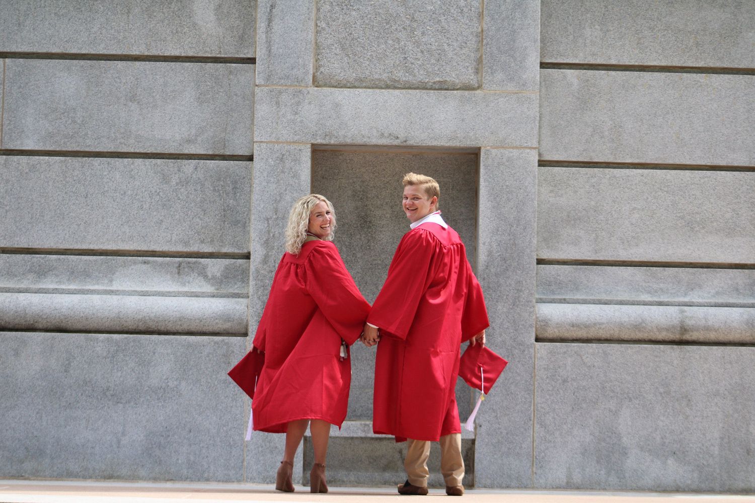 Two graduates in red robes hold hands in front of stone building.