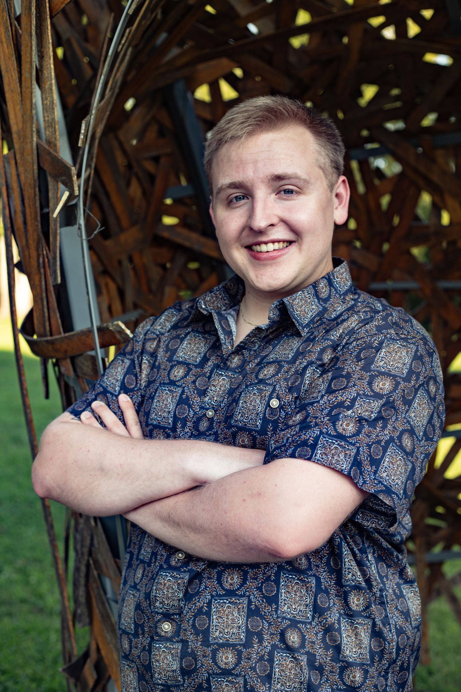 A person in a blue patterned shirt poses with crossed arms against a natural outdoor background.