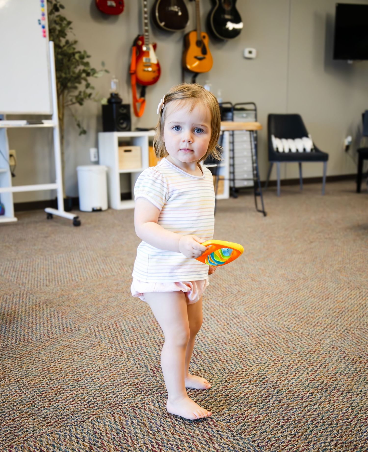Small toddler in striped shirt and diaper stands and plays with toys in a room with guitars hanging on the wall.