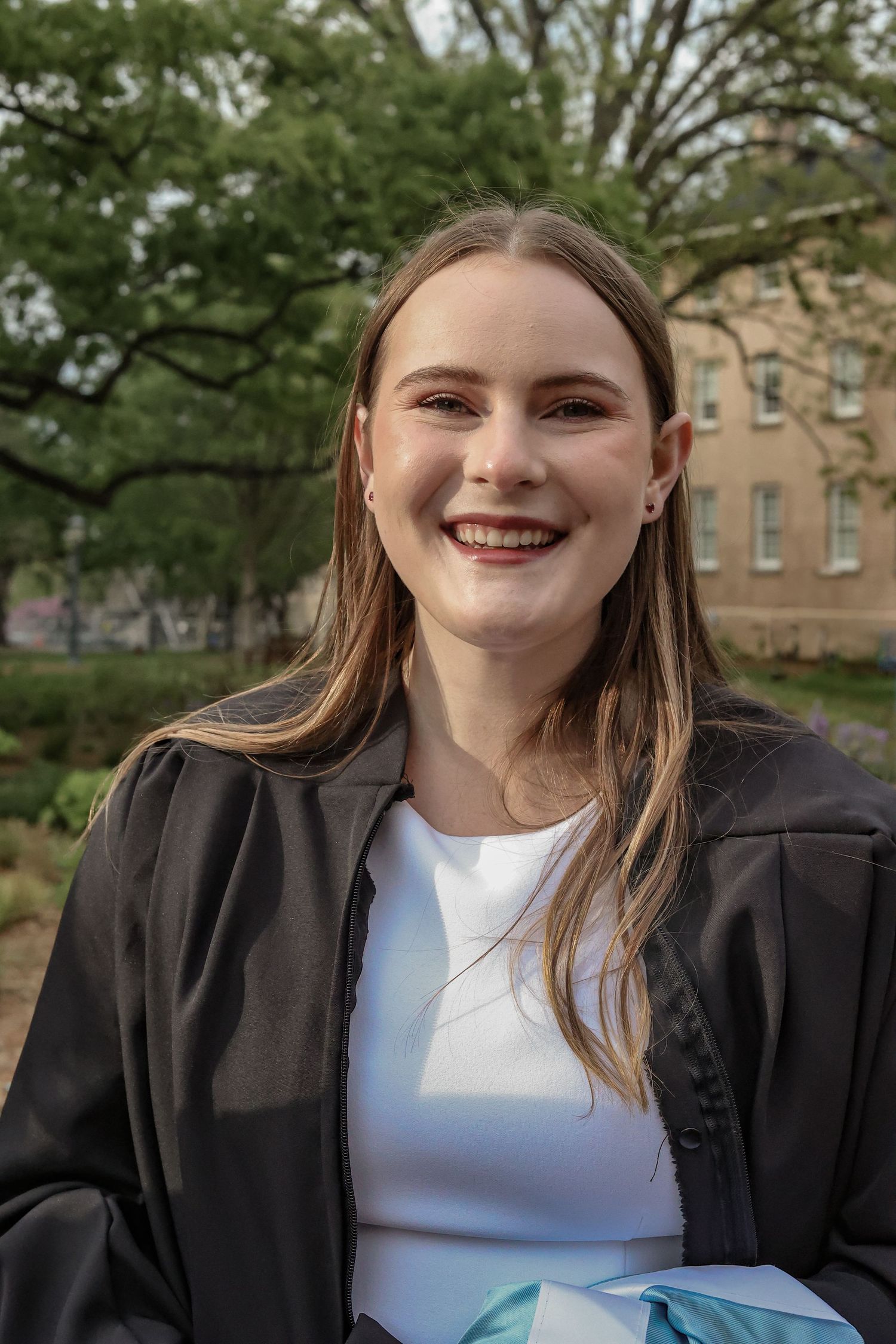 Graduate in a black cap and gown smiling against an outdoor campus backdrop.