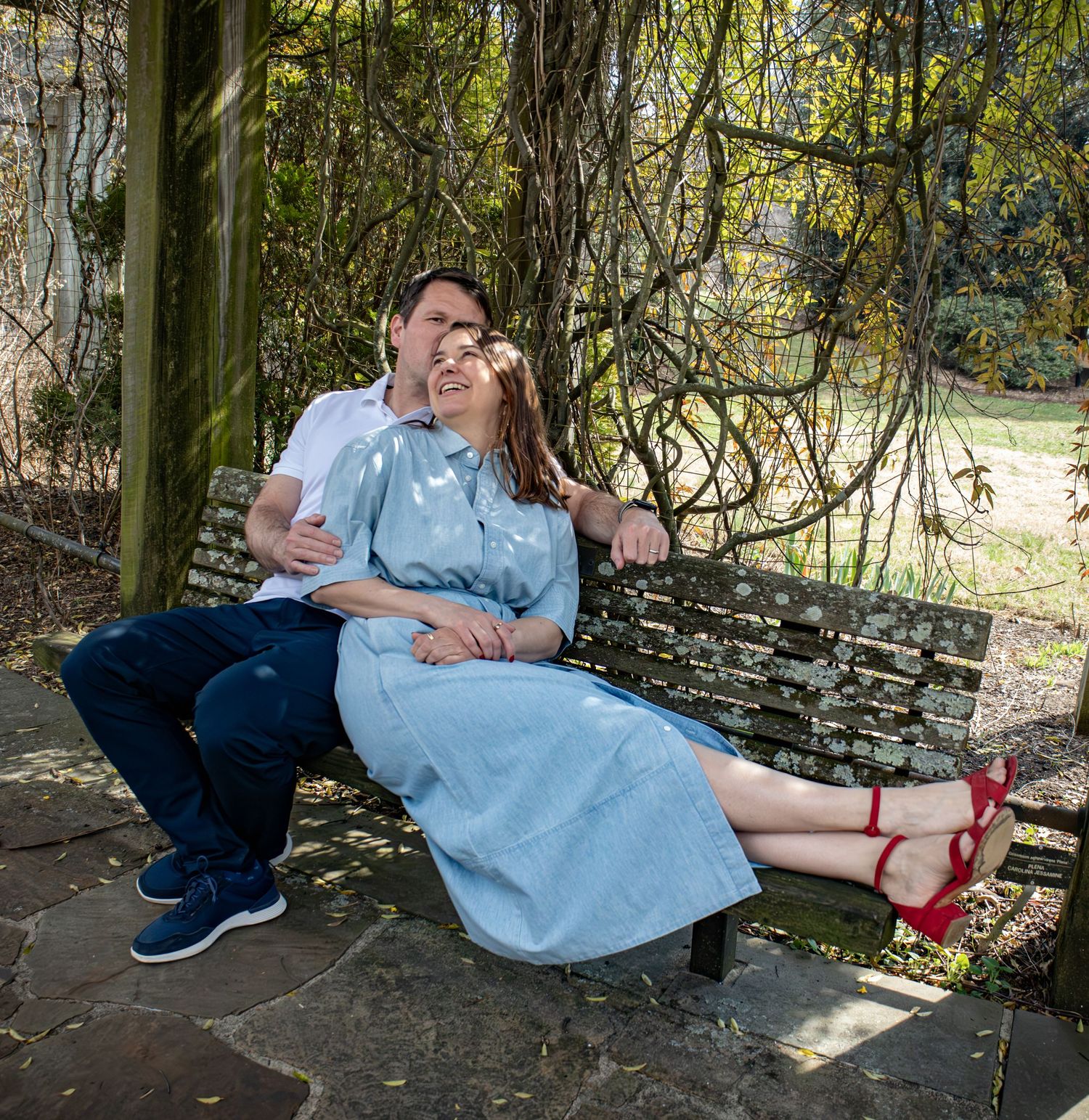 A couple relaxes on a stone bench under hanging vines in a garden setting.