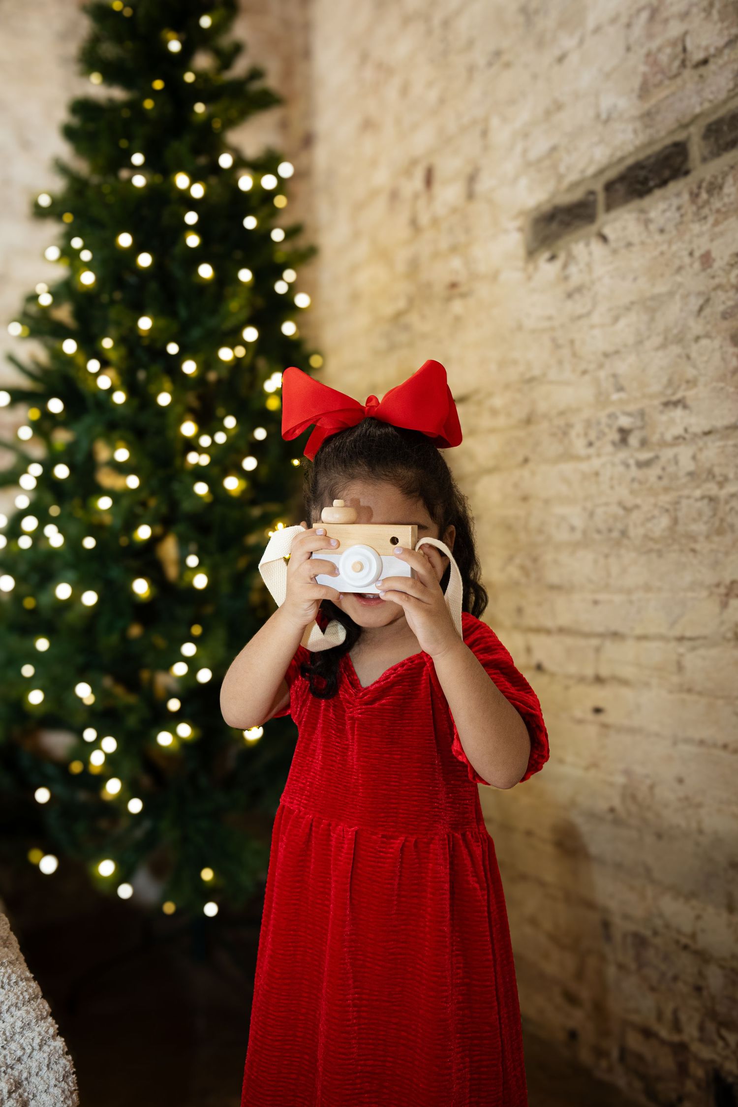 Child in red dress with bow holding toy camera near Christmas tree.