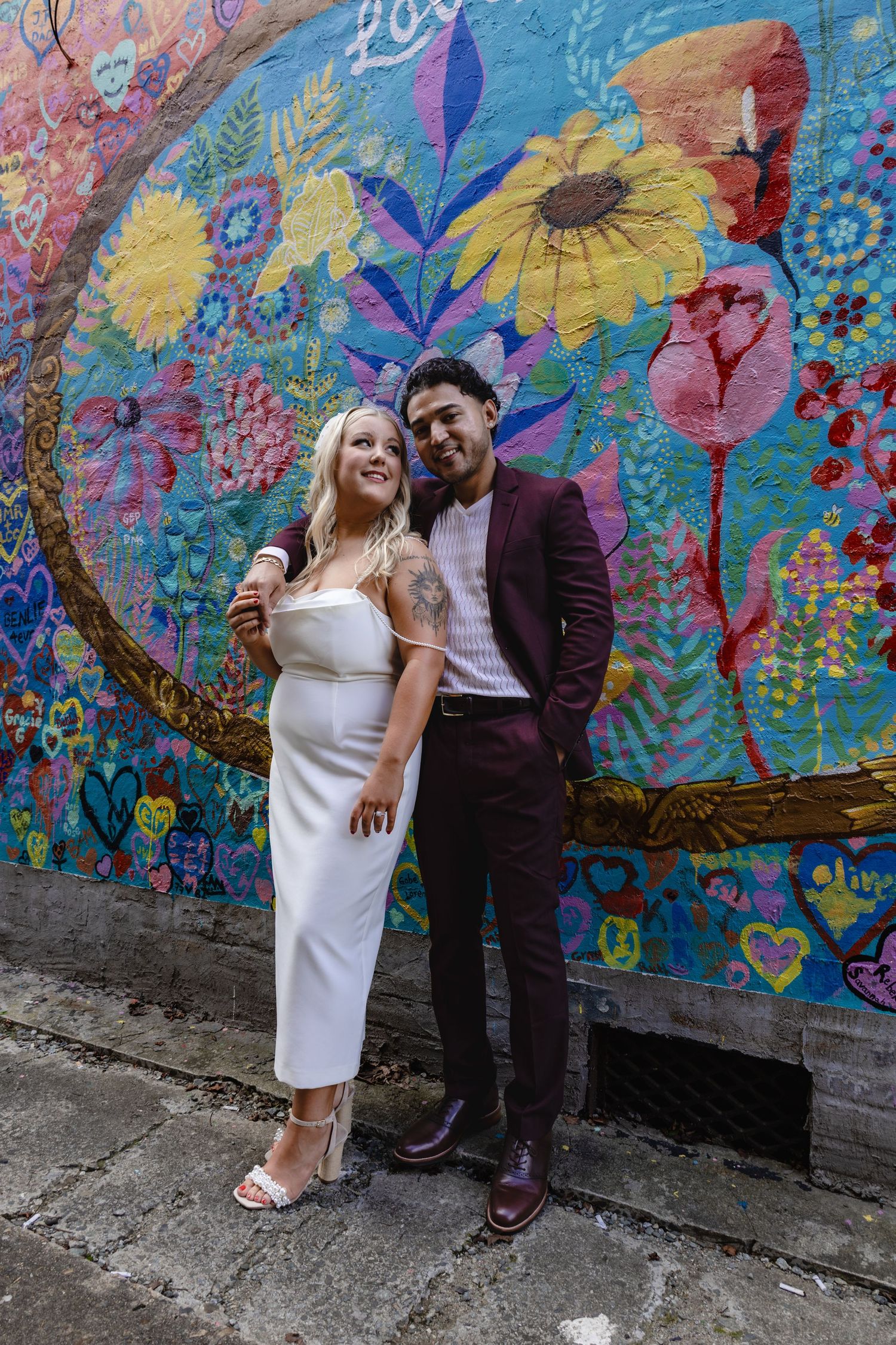 A couple in formal attire poses against a bright floral mural background.