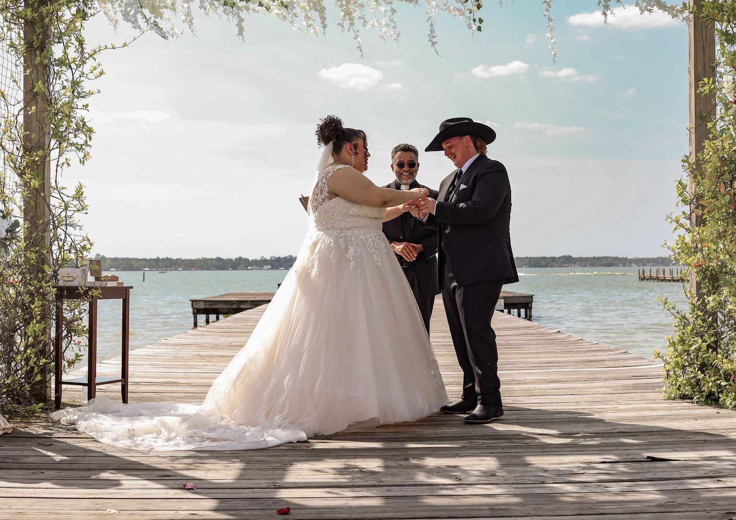 Wedding ceremony taking place at sunset on a scenic waterfront pier with trees creating a romantic archway.