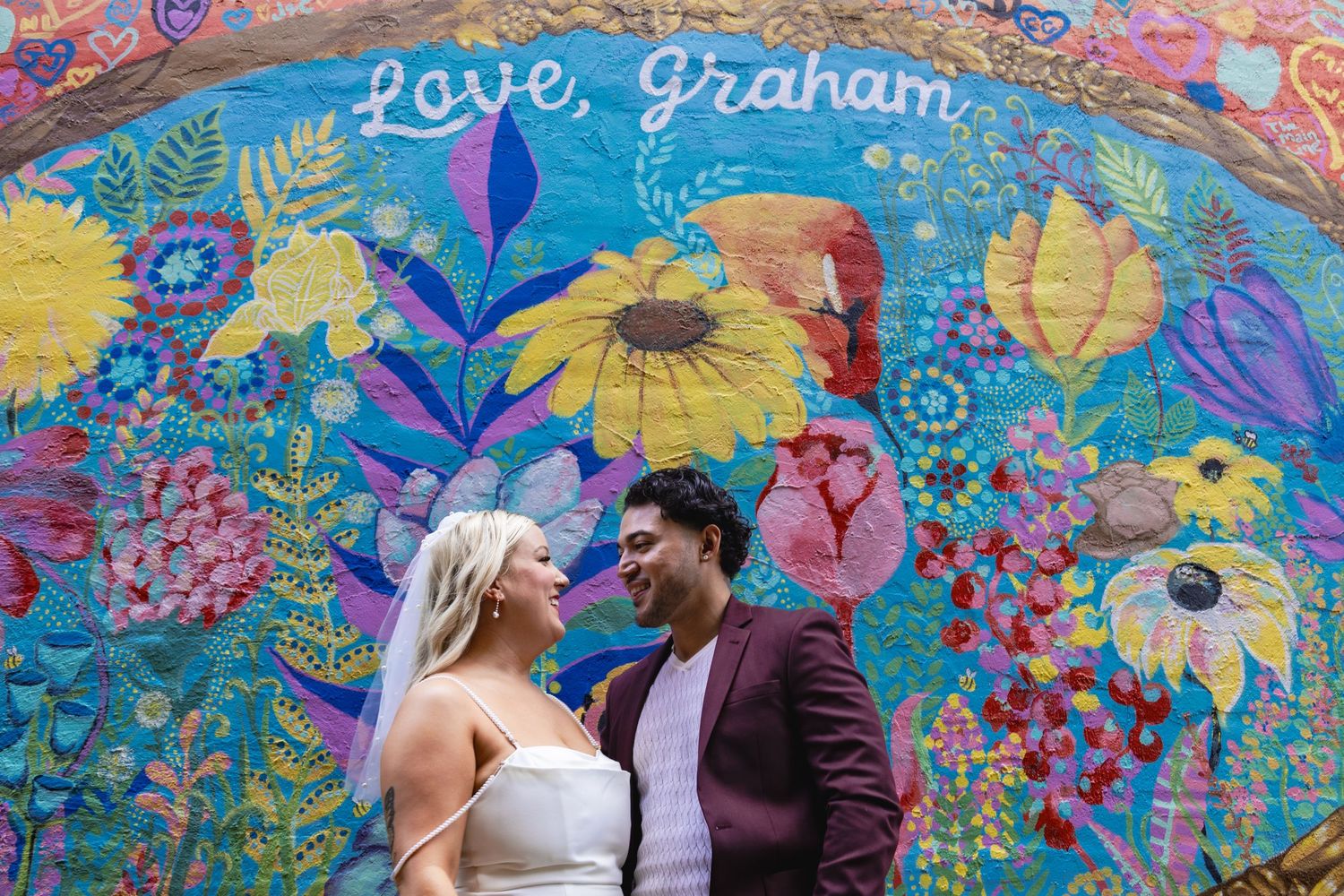 A couple poses in front of a vibrant floral mural with 'Love, Graham' text.