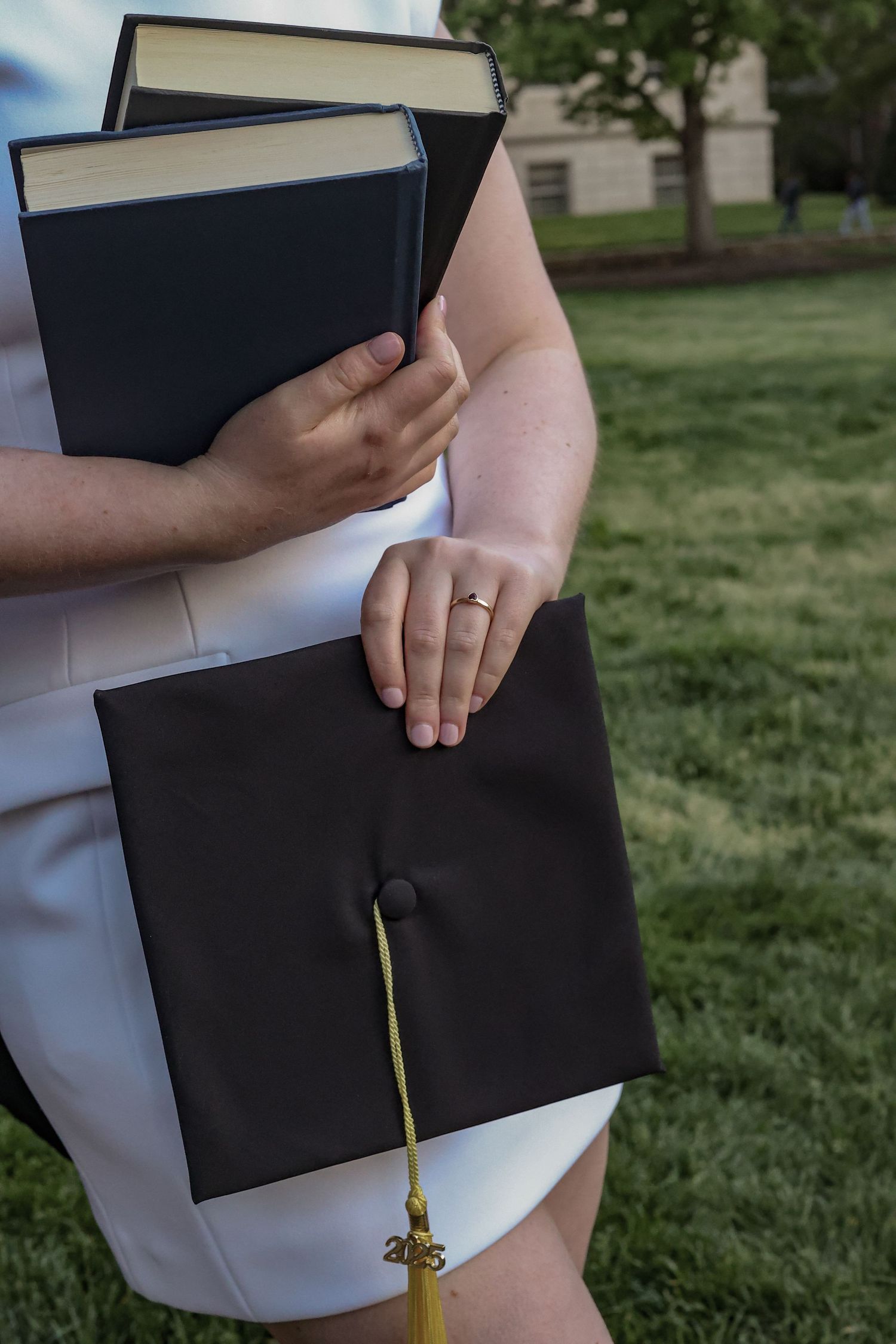 Close-up of graduate holding books and graduation cap showing engagement ring.