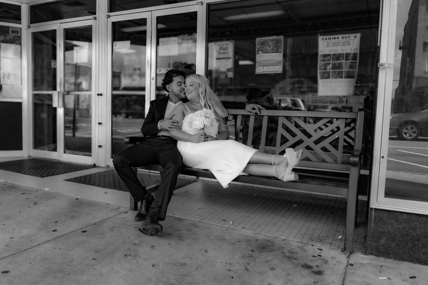 Black and white photo of a couple sitting together on a wooden bench outside a storefront.