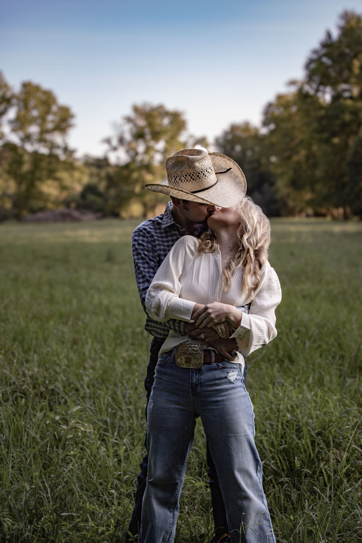 A couple in western wear embrace in a sunlit meadow with trees in the background.