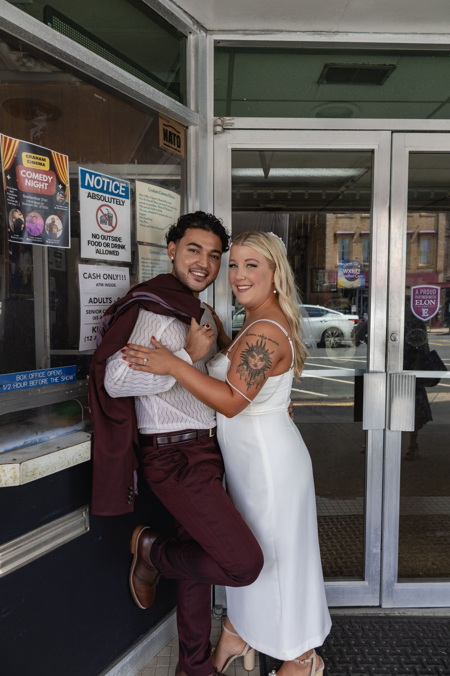 A couple in wedding attire playfully poses outside a shop entrance.