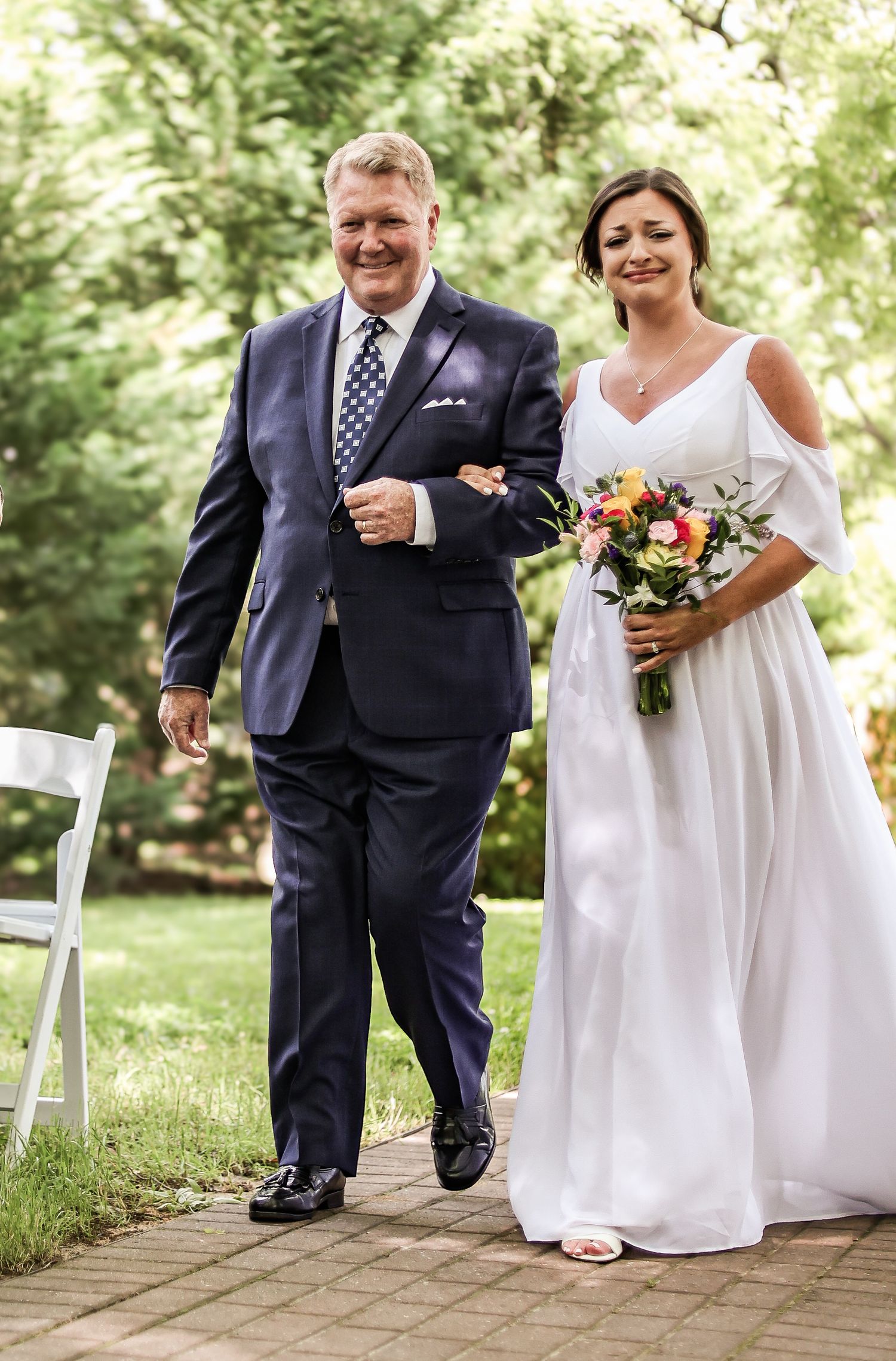 White wedding ceremony in outdoor garden setting with bride in flowing dress holding colorful bouquet walking down path.