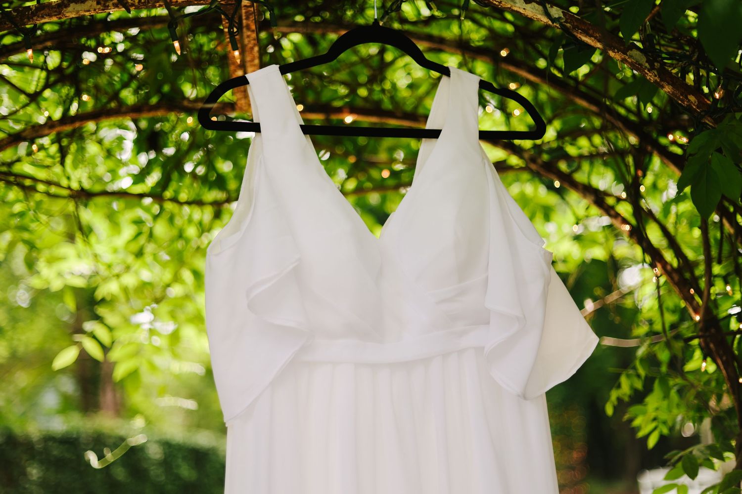 A white flowy wedding dress hangs from tree branches in a lush green garden setting.