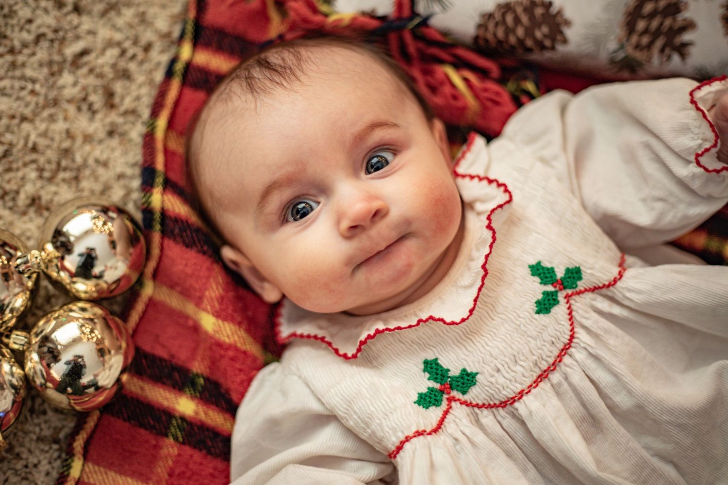 A baby wearing a white and red holiday outfit lies on a festive red blanket with pinecones in the background.