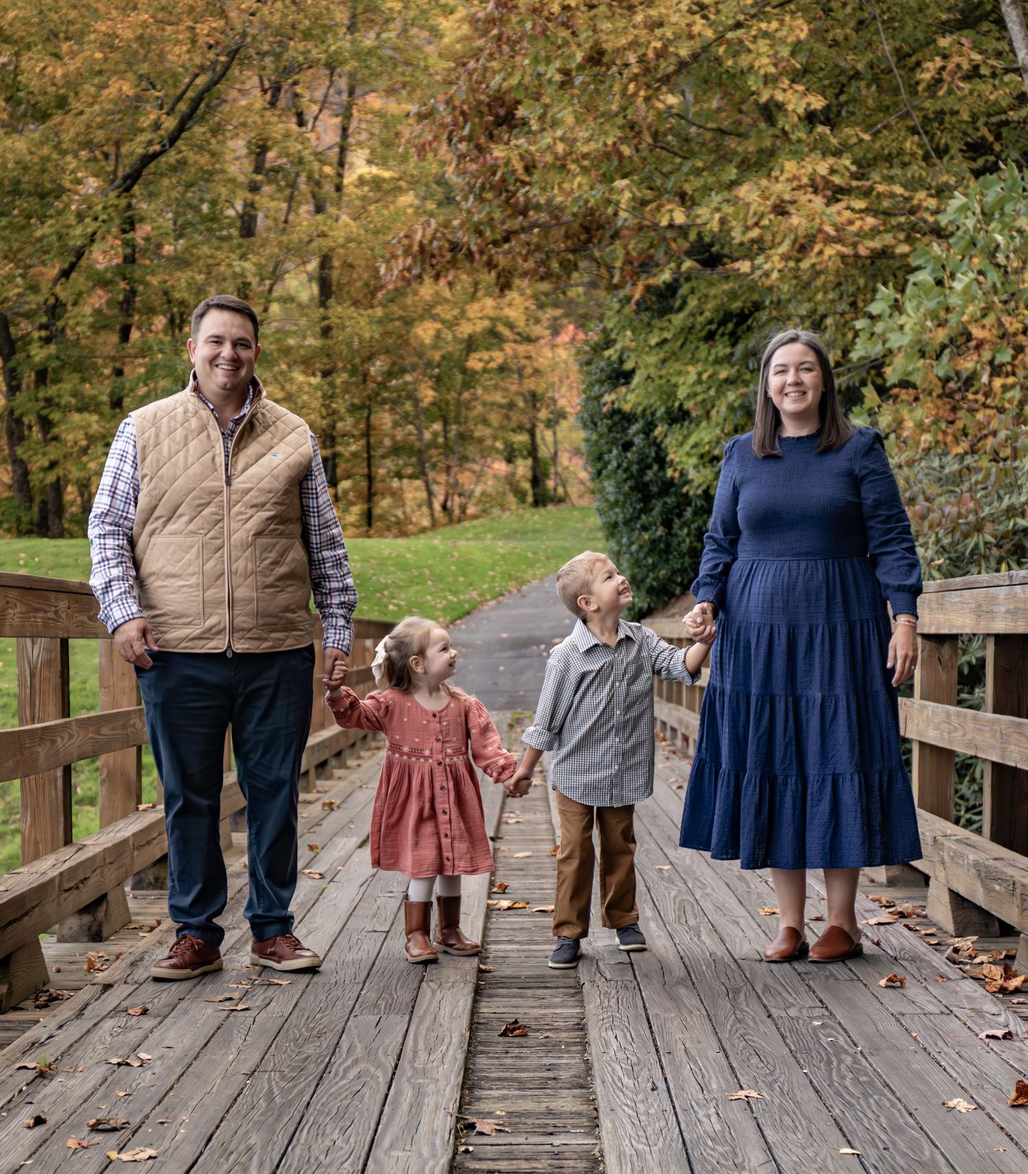 A family stands together on a wooden bridge surrounded by autumn trees with golden foliage.