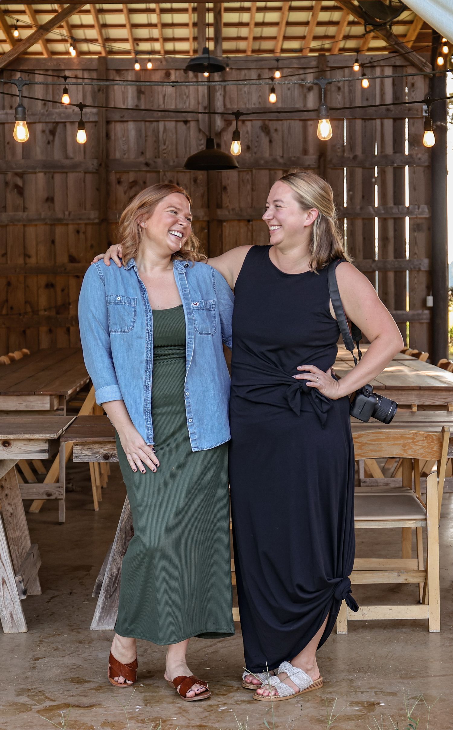 Two people in casual dresses share a laugh while sitting together in a rustic barn setting with string lights overhead.