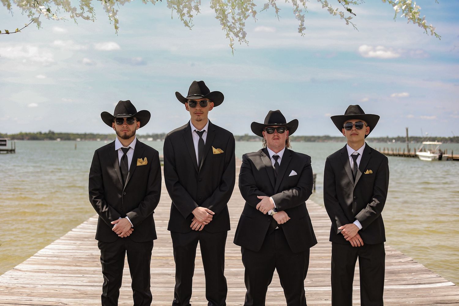 Groomsmen in matching black suits and cowboy hats posing together on a dock with water in background.