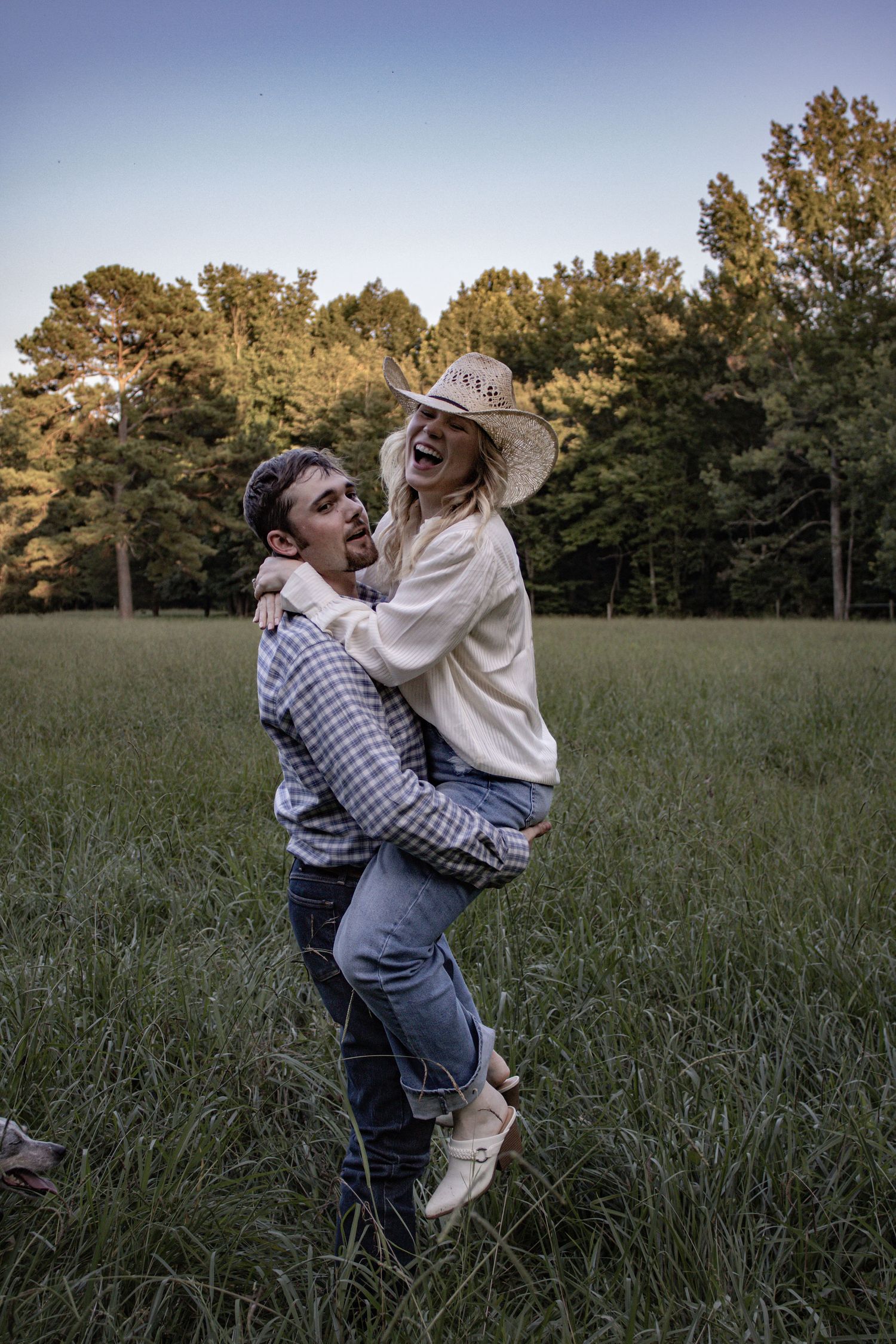 A couple shares a playful moment in a field at sunset as one person lifts the other wearing a cowboy hat.