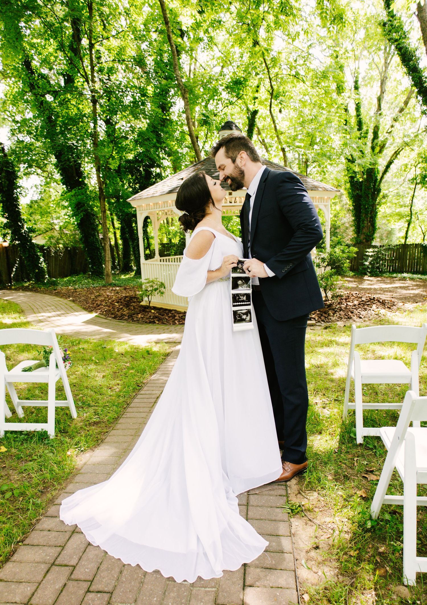 A couple in wedding attire share a tender moment on a stone path surrounded by greenery and white chairs at an outdoor venue.