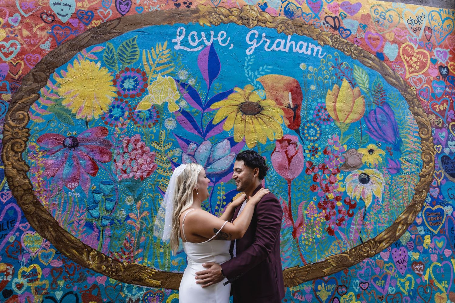 A couple embraces in front of a vibrant blue mural with yellow sunflowers.